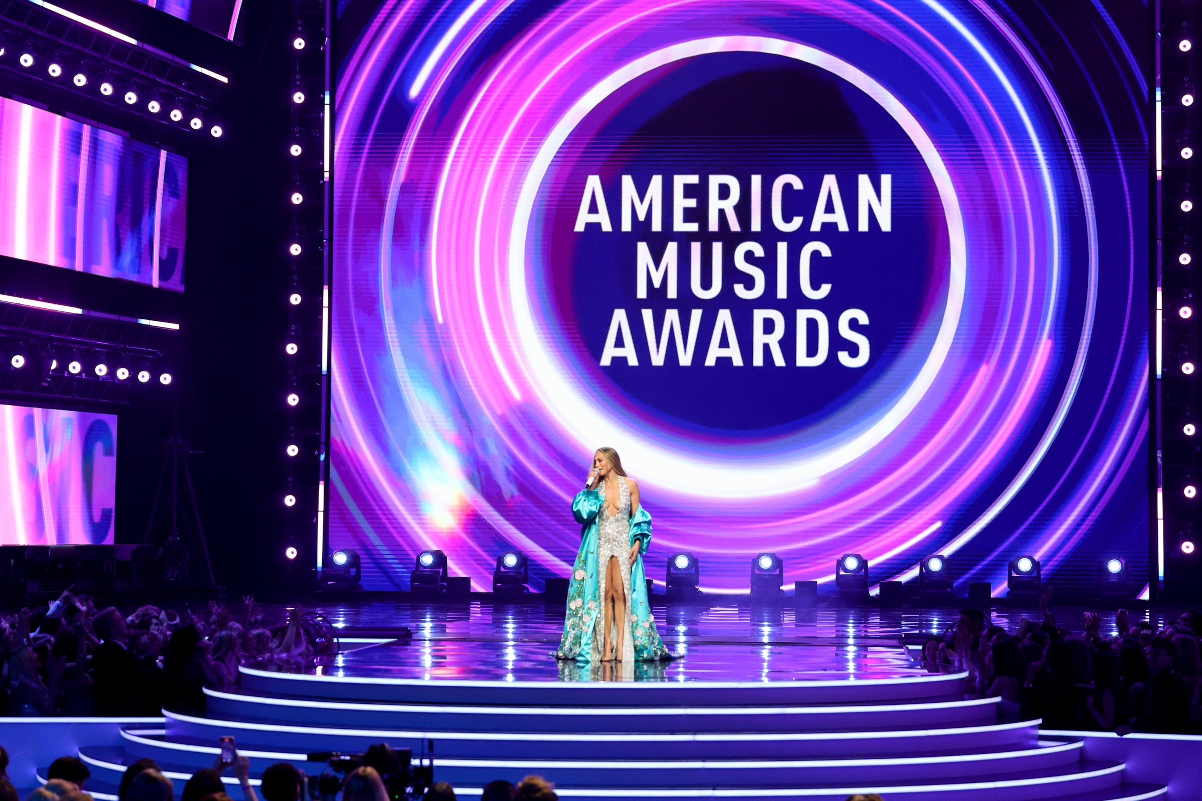 In a wide shot of the dazzling stage, the 55-year-old songstress takes center spotlight at the American Music Awards, framed by swirling neon visuals as the audience looks on.