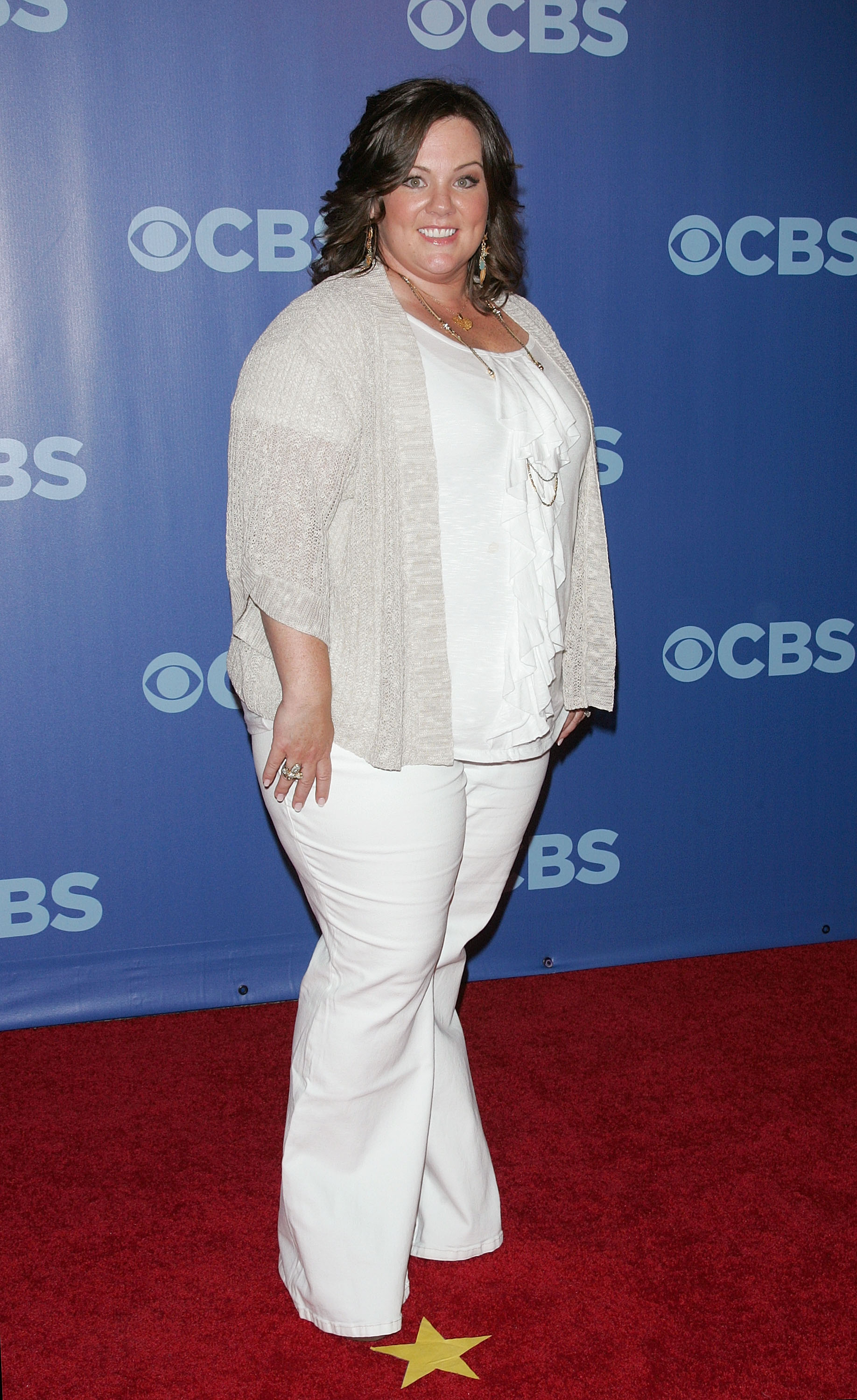 Melissa McCarthy appears smiling on the red carpet, dressed in soft neutral layers paired with white pants that brighten the look. Against the bold blue backdrop, her relaxed outfit and confident pose create a warm, welcoming presence.