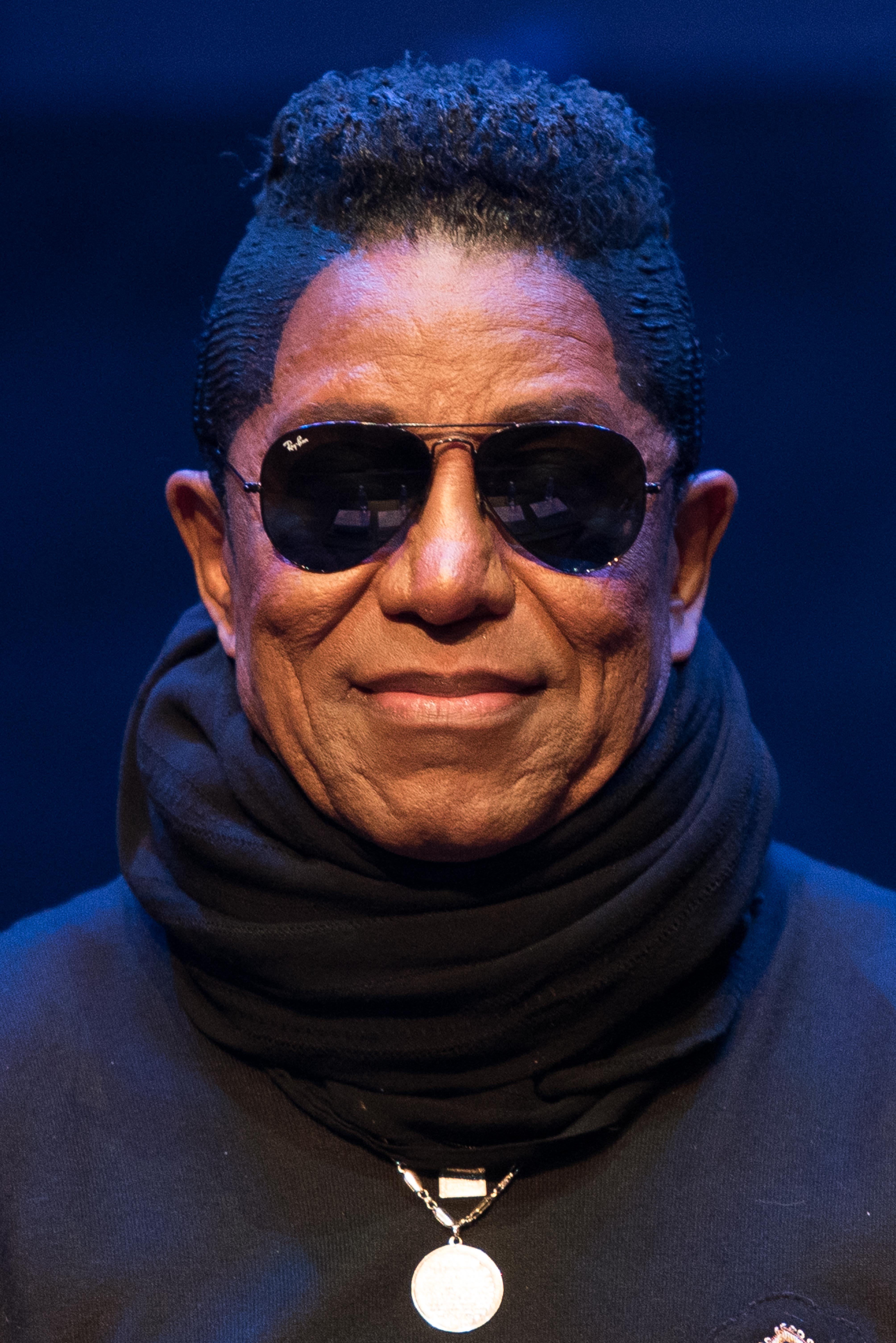Jermaine Jackson appears at the Forever Michael Jackson show in Madrid, dressed in an all-black ensemble with a high scarf and dark sunglasses. The sleek, layered styling and confident stance give him a modern, almost enigmatic stage presence.
