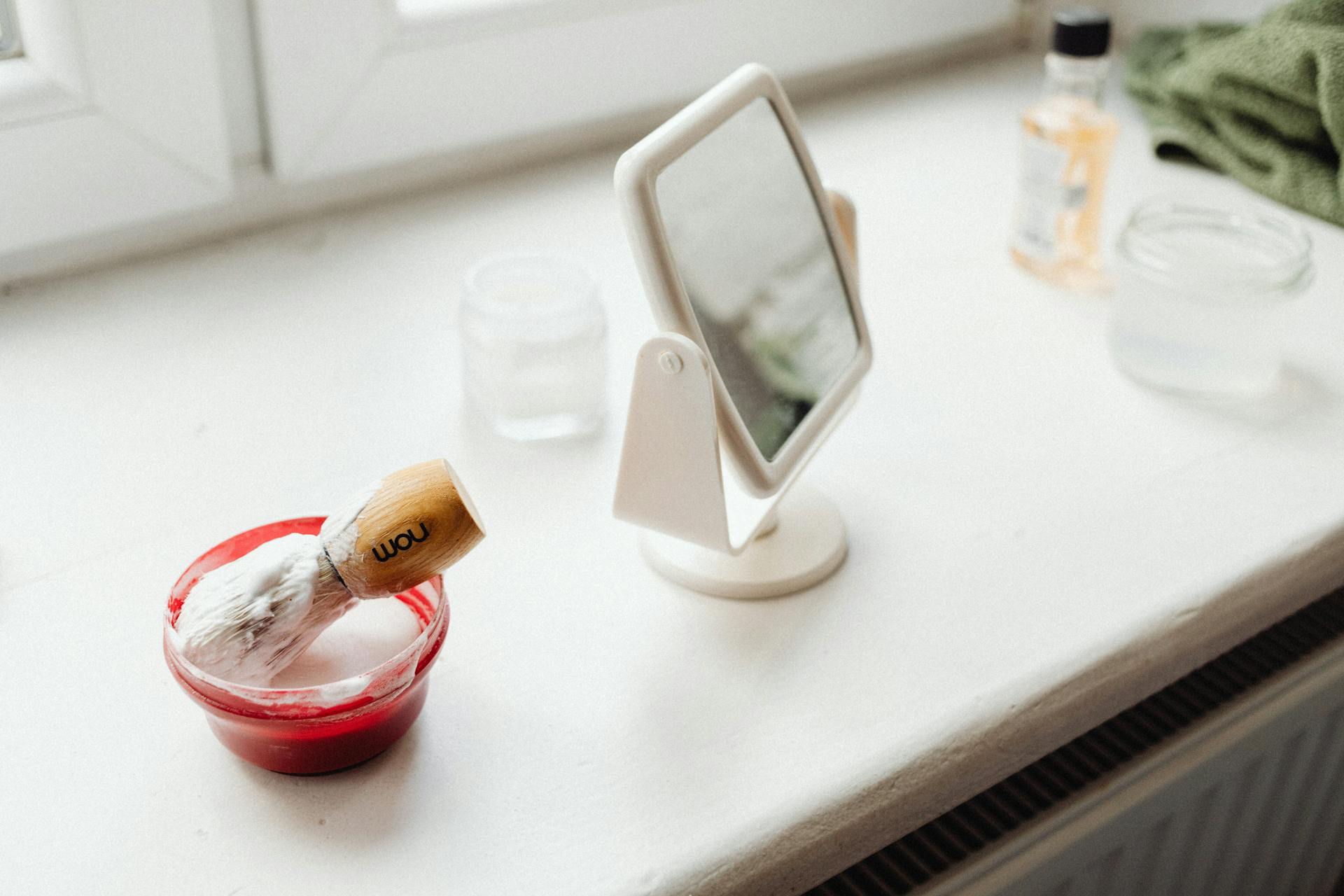 A close-up of a mirror and a container with shaving cream and a shaving brush on a windowsill | Source: Pexels