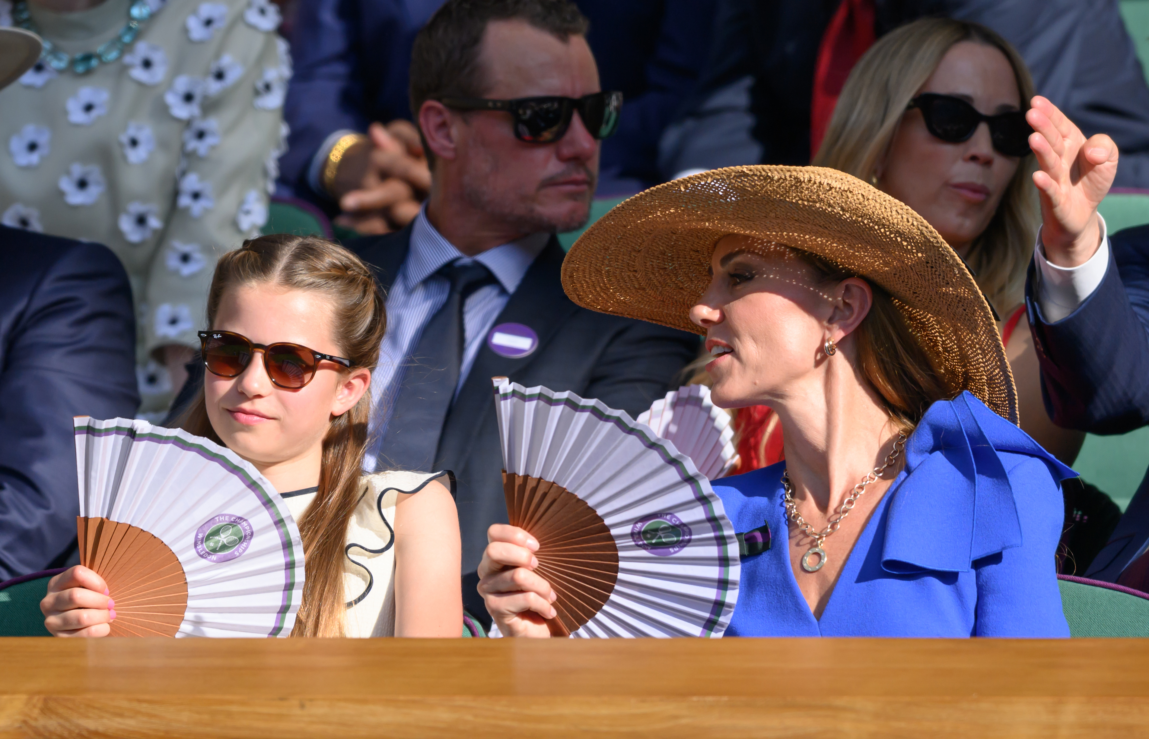 Princess Catherine leans in to speak with Princess Charlotte as they sit together in the Royal Box. Both hold matching fans while shielding themselves from the sun in the packed Wimbledon stands.