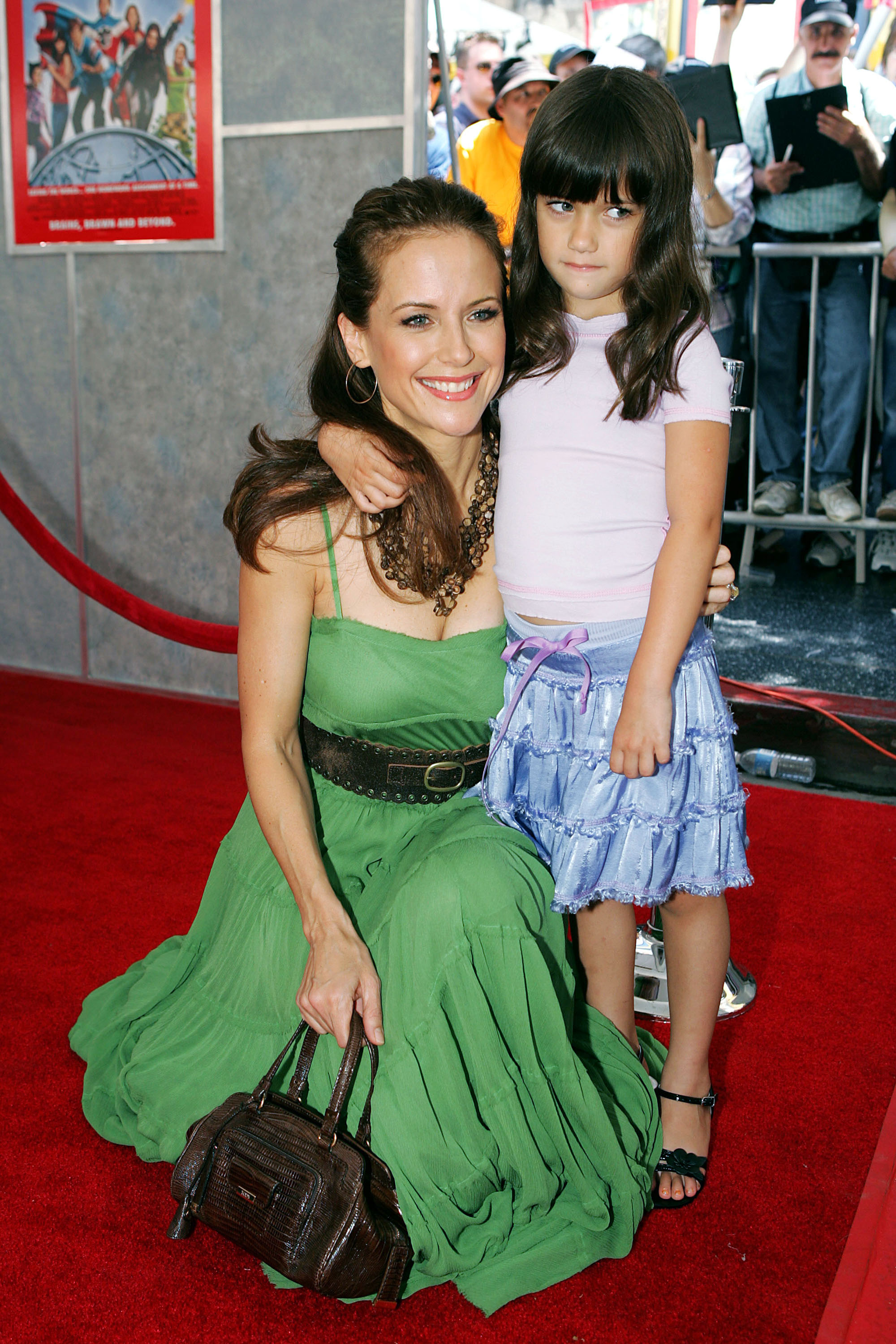 Kelly Preston kneels beside Ella Bleu Travolta on the red carpet, smiling in a flowing green dress while Ella wears a pale pink top and blue skirt. They attended the 2005 premiere of "Sky High" in Hollywood.