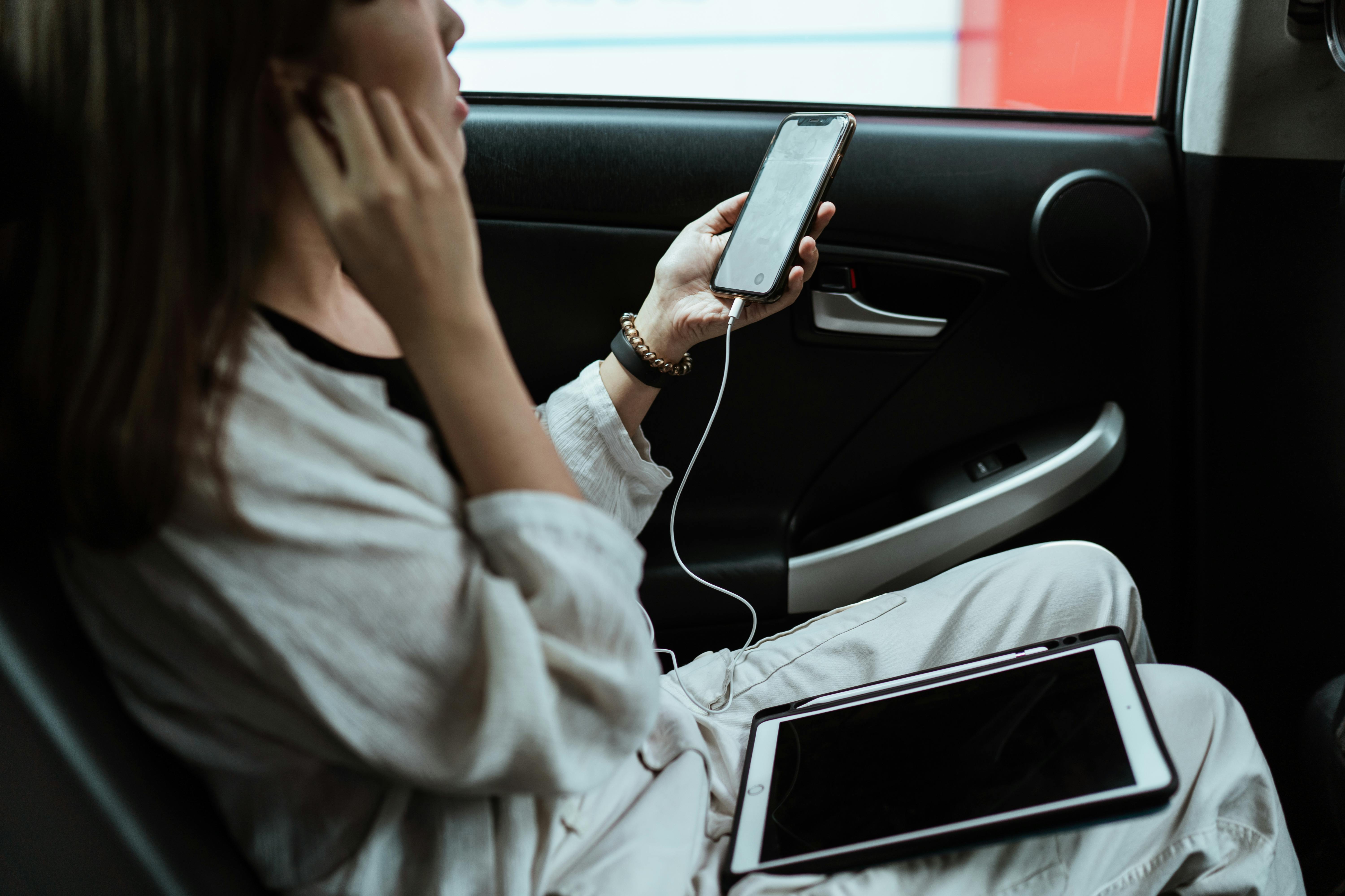 A woman sitting in a car holding a phone, wearing headphones and a laptop on her lap | Source: Pexels