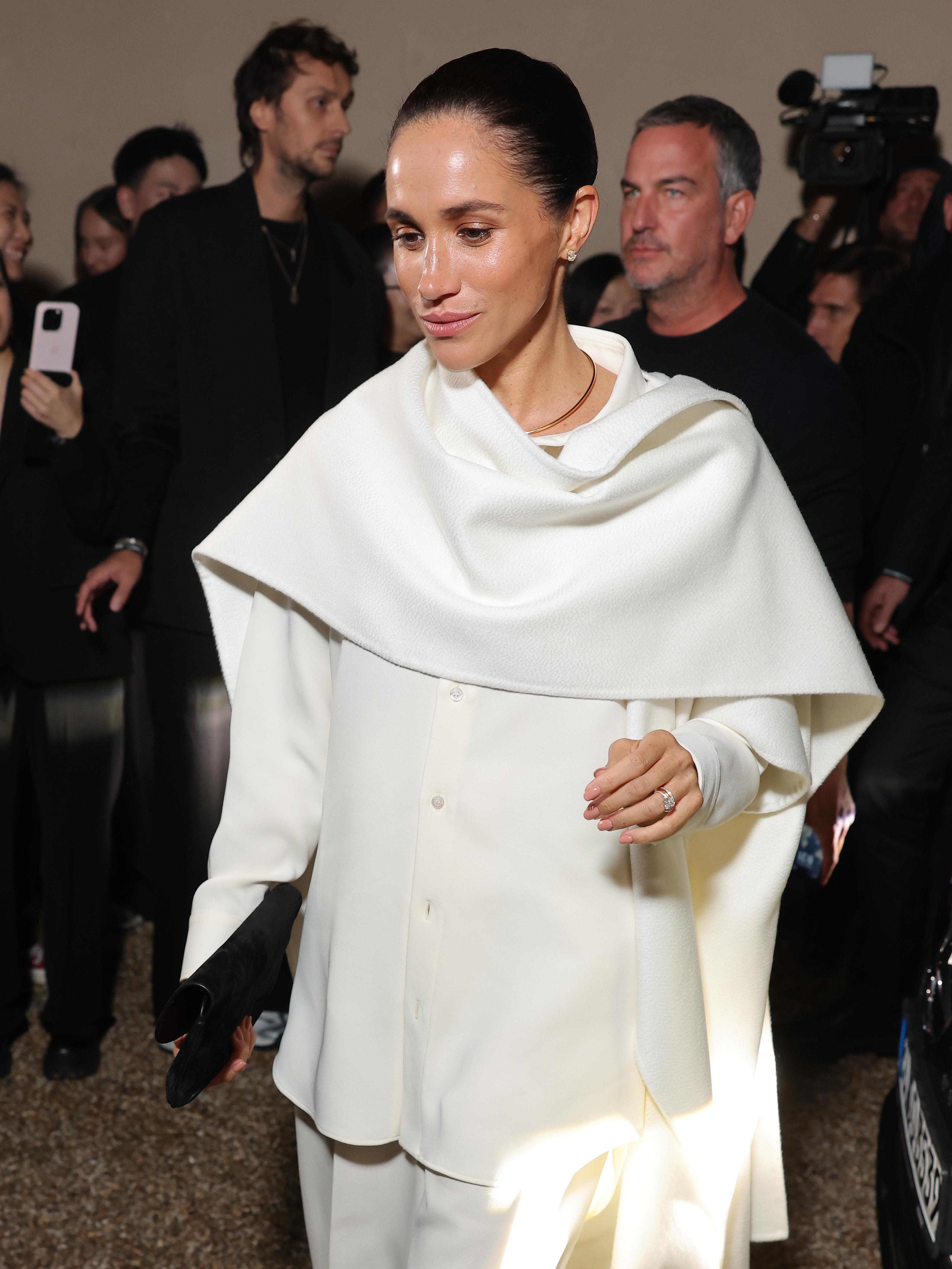 Meghan Markle attends the Womenswear Spring/Summer 2026 show in Paris, dressed in an elegant white cape layered over a matching shirt, her hair styled in a sleek bun as she smiles for the cameras.