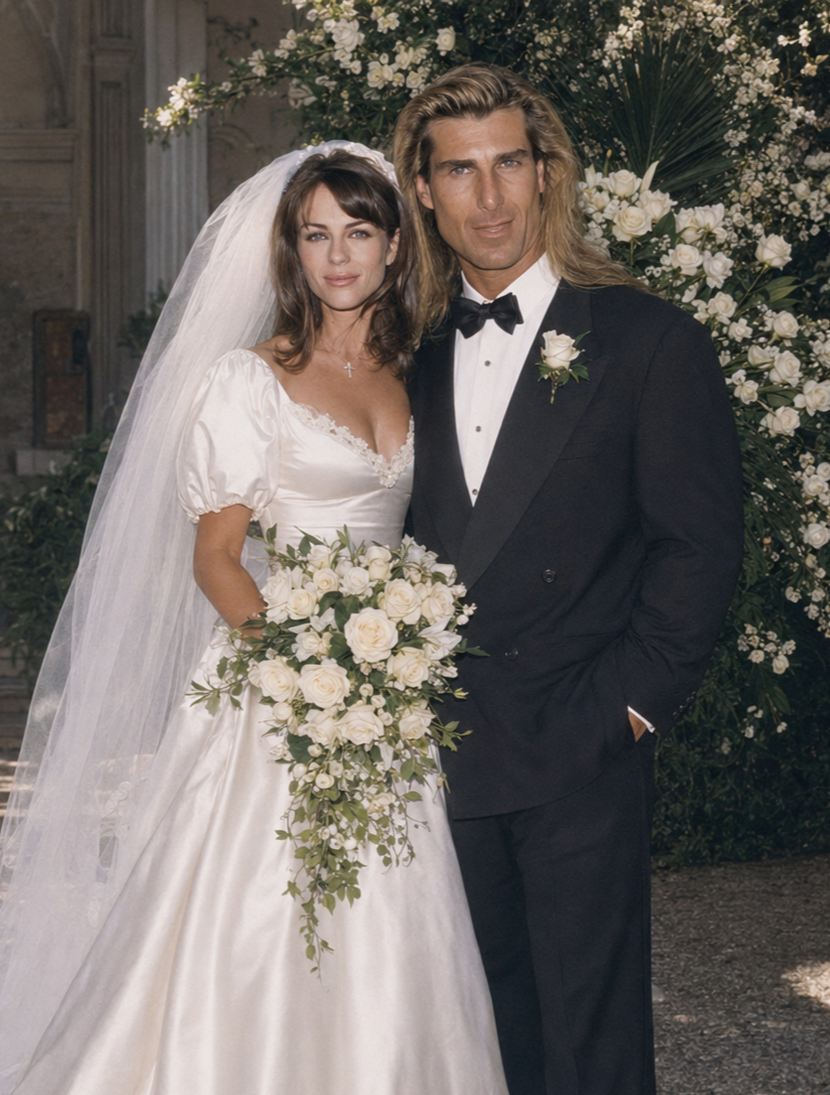 This AI creation portrays Fabio in a classic double-breasted black tuxedo posing next to Elizabeth Hurley in an elegant white bridal gown with puffed sleeves and a veil, holding a lush cascading bouquet. | Source: ChatGPT