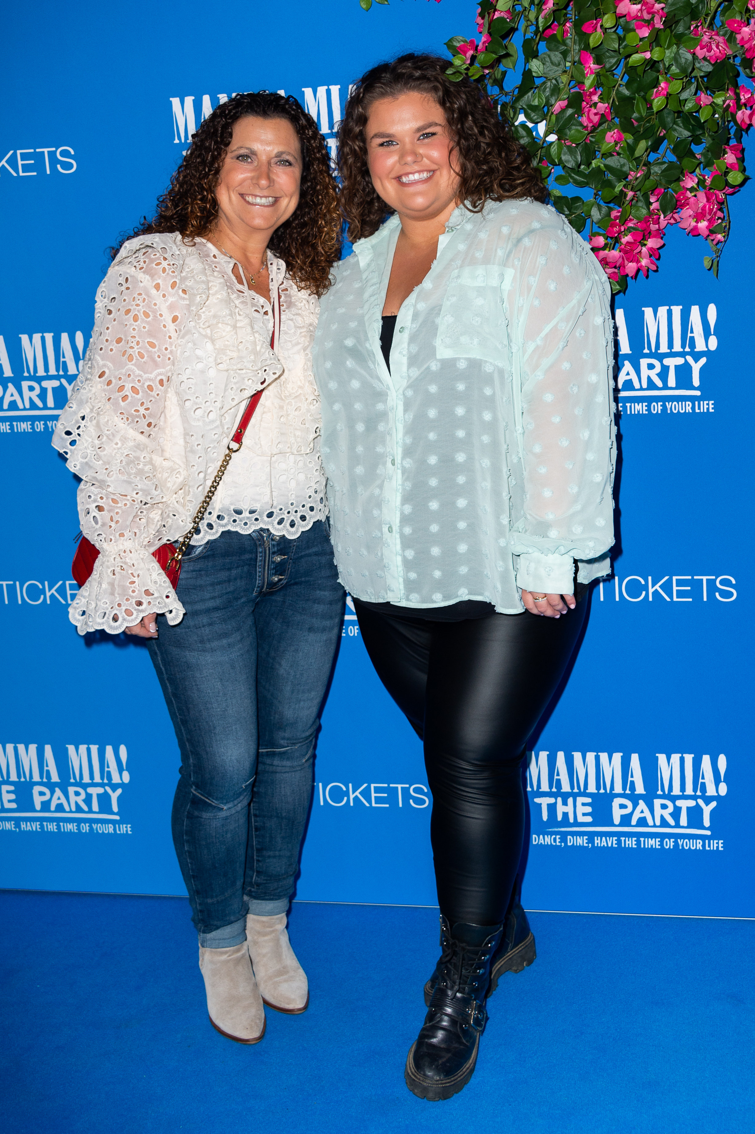 Tapper and her mother Nikki lean in close together on the blue carpet at the "Mamma Mia!" The Party gala at The O2 Arena, London, September 2023, both smiling broadly beneath a cascade of pink floral decorations overhead.