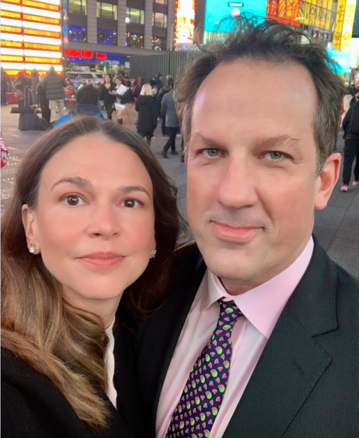Sutton Foster stands beside Ted Griffin for a city selfie surrounded by bright lights and crowds. While Sutton keeps it polished in a dark coat and soft glam makeup, Ted wears a suit with a colorful patterned tie. | Source: Instagram/suttonlenore