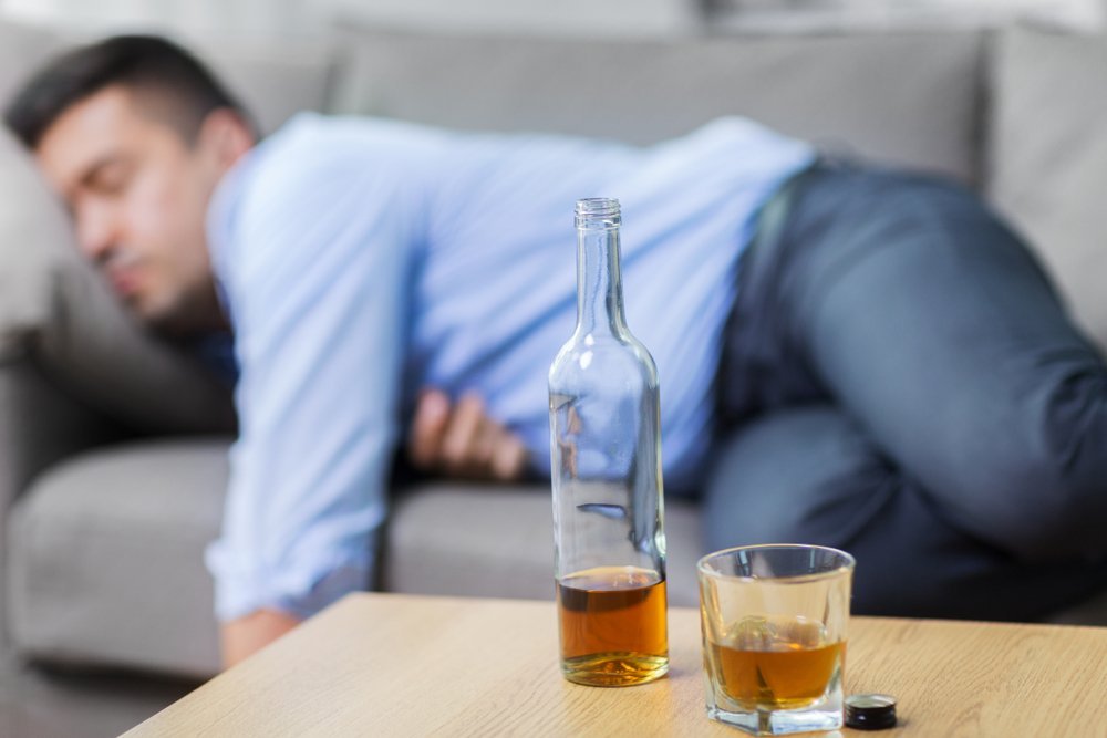 A photo of a sleeping drunk man with a bottle and glass of whiskey on table. | Photo: Shutterstock