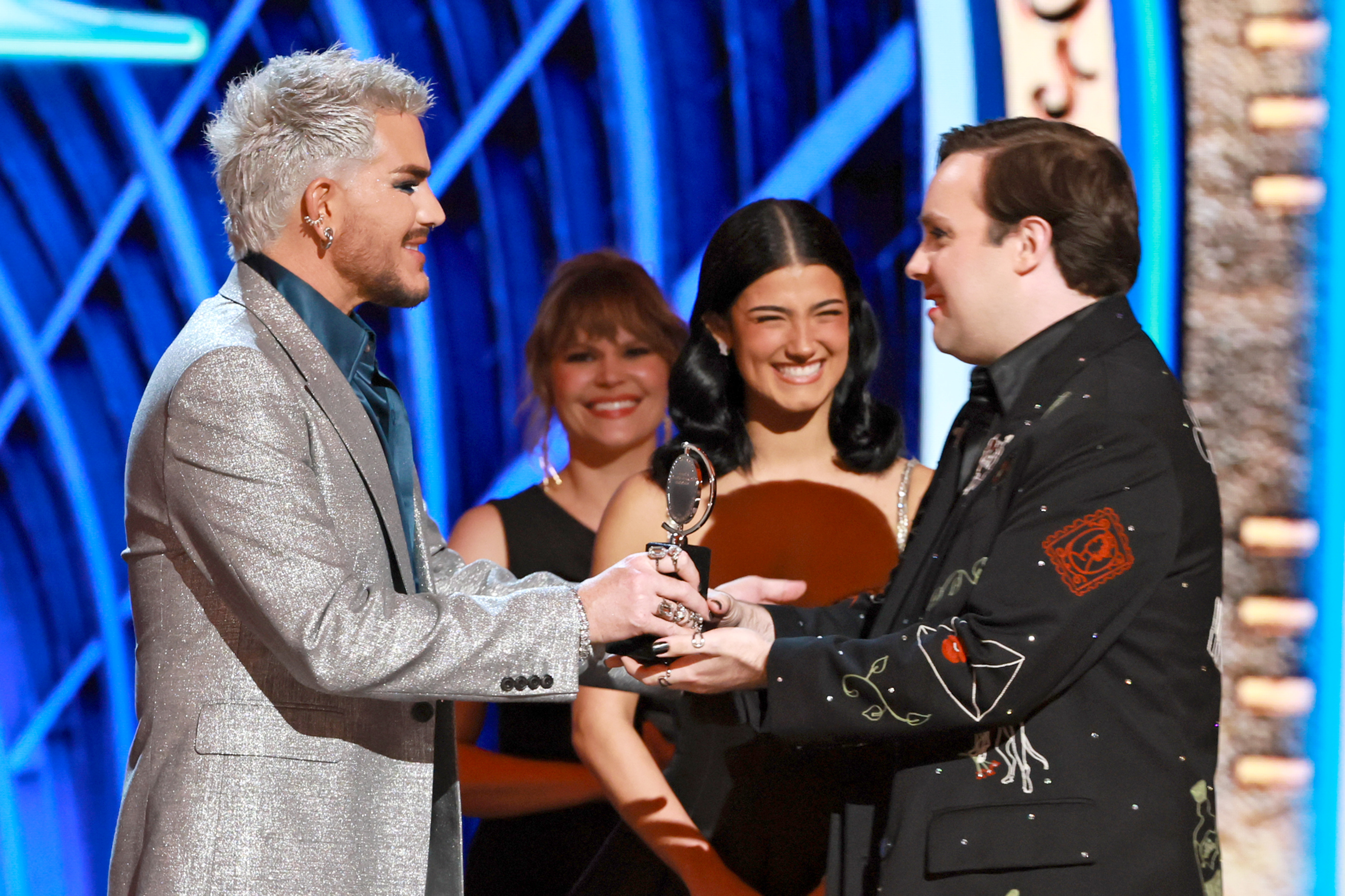 Adam Lambert hands a Tony statuette to Jak Malone as Charli D'Amelio and other presenters look on. Lambert's poised posture and Malone's emotional reaction capture a heartfelt exchange at a high point in the night's honors.