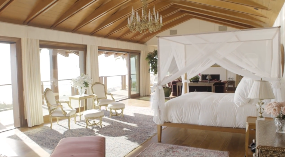 Sliding glass doors line the wall of the primary suite, filling the room with ocean light and providing immediate access to the upper balcony. A white-draped canopy bed rests under high wooden ceiling beams, while a classic brass chandelier and a gilded seating set add sophisticated charm to the layout. | Source: YouTube/Architectural Digest