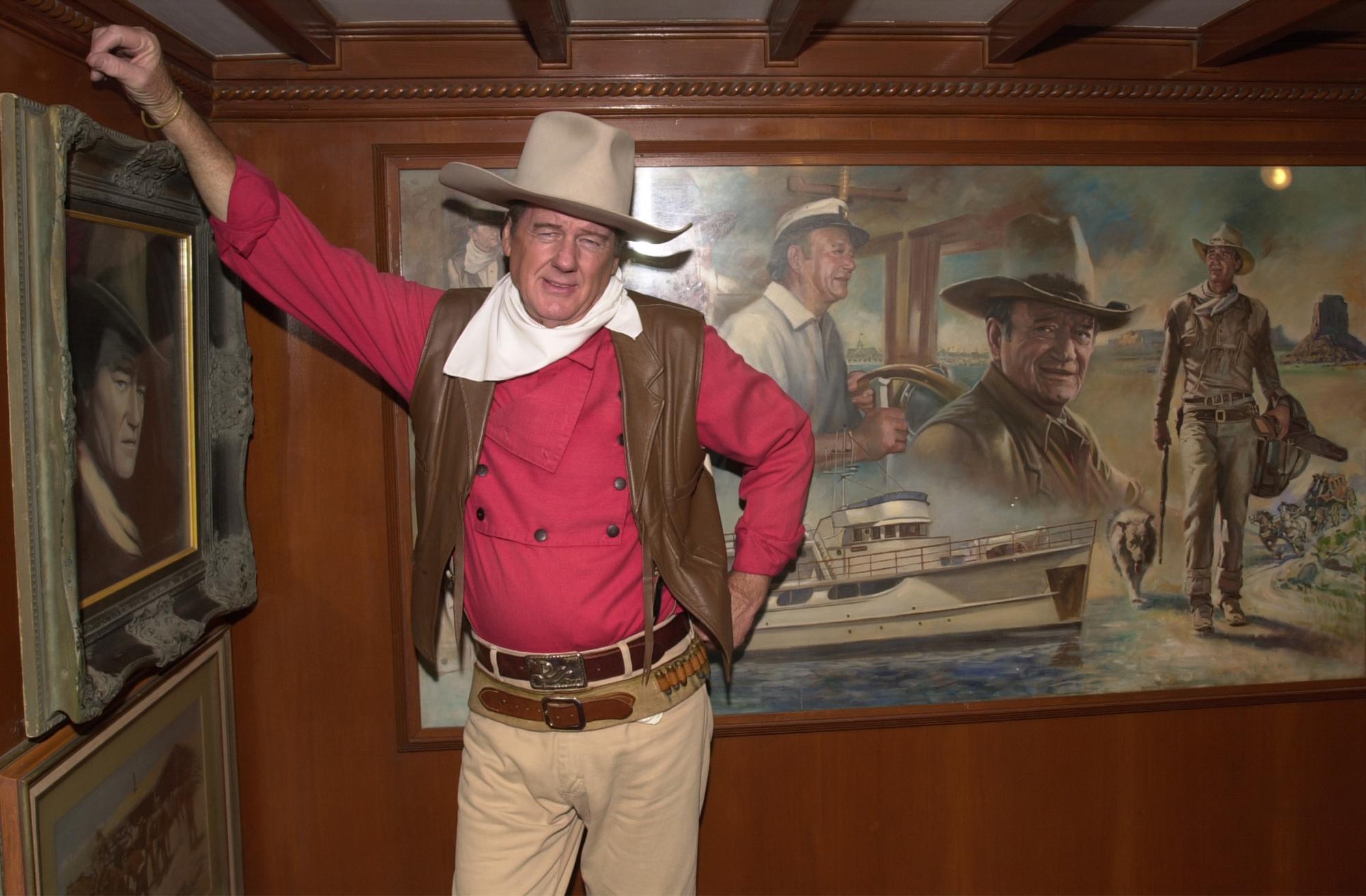 John Wayne impersonator Ermal Williamson stands in front of portraits of John Wayne at the Fall 2000 Book Bash on September 20, 2000 in Newport Beach, California. | Source: Getty Images
