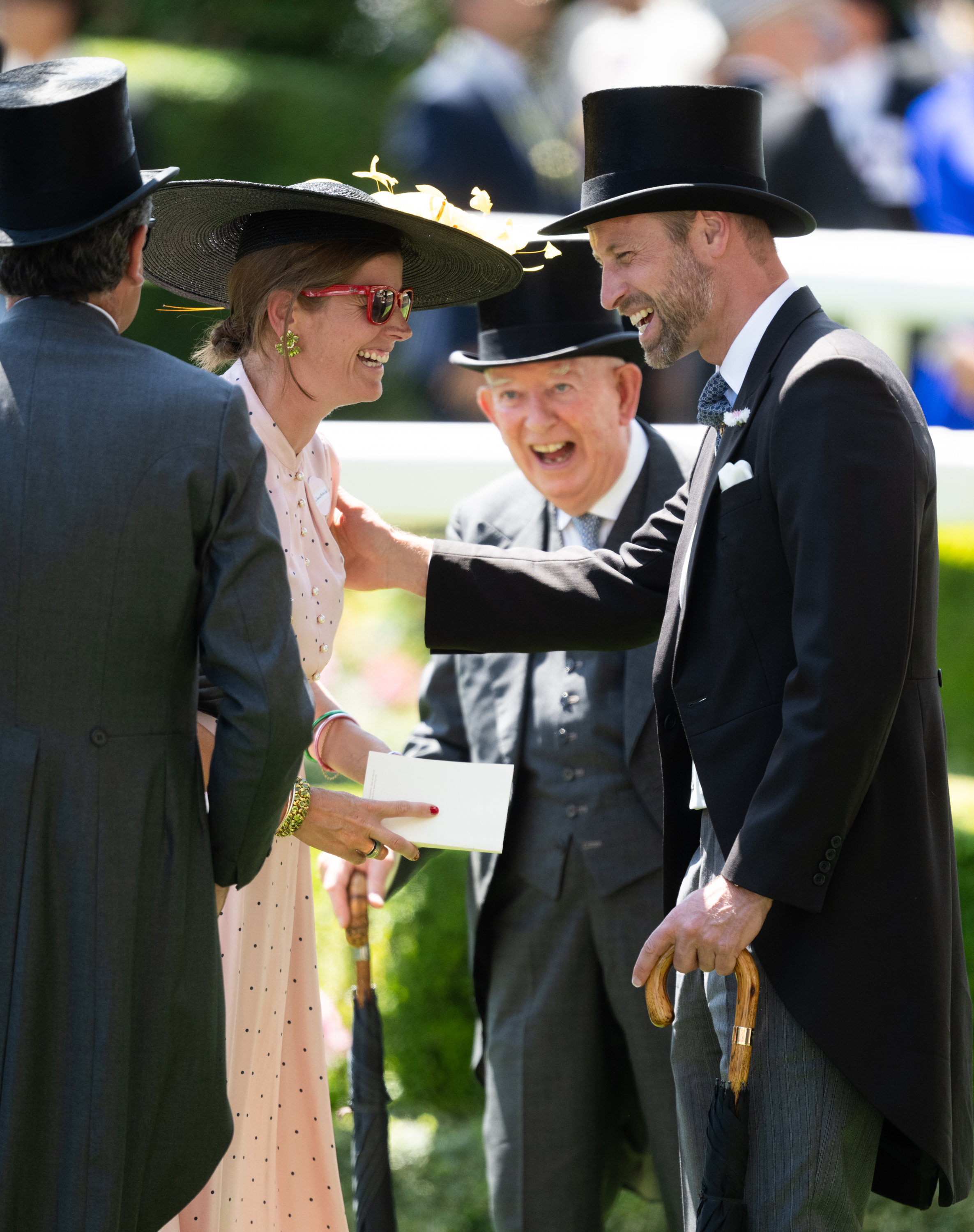 Prince William shares a lighthearted exchange with guests at Royal Ascot, smiling and laughing as he greets a woman in a wide-brimmed black hat and polka-dot dress on the racecourse lawn.