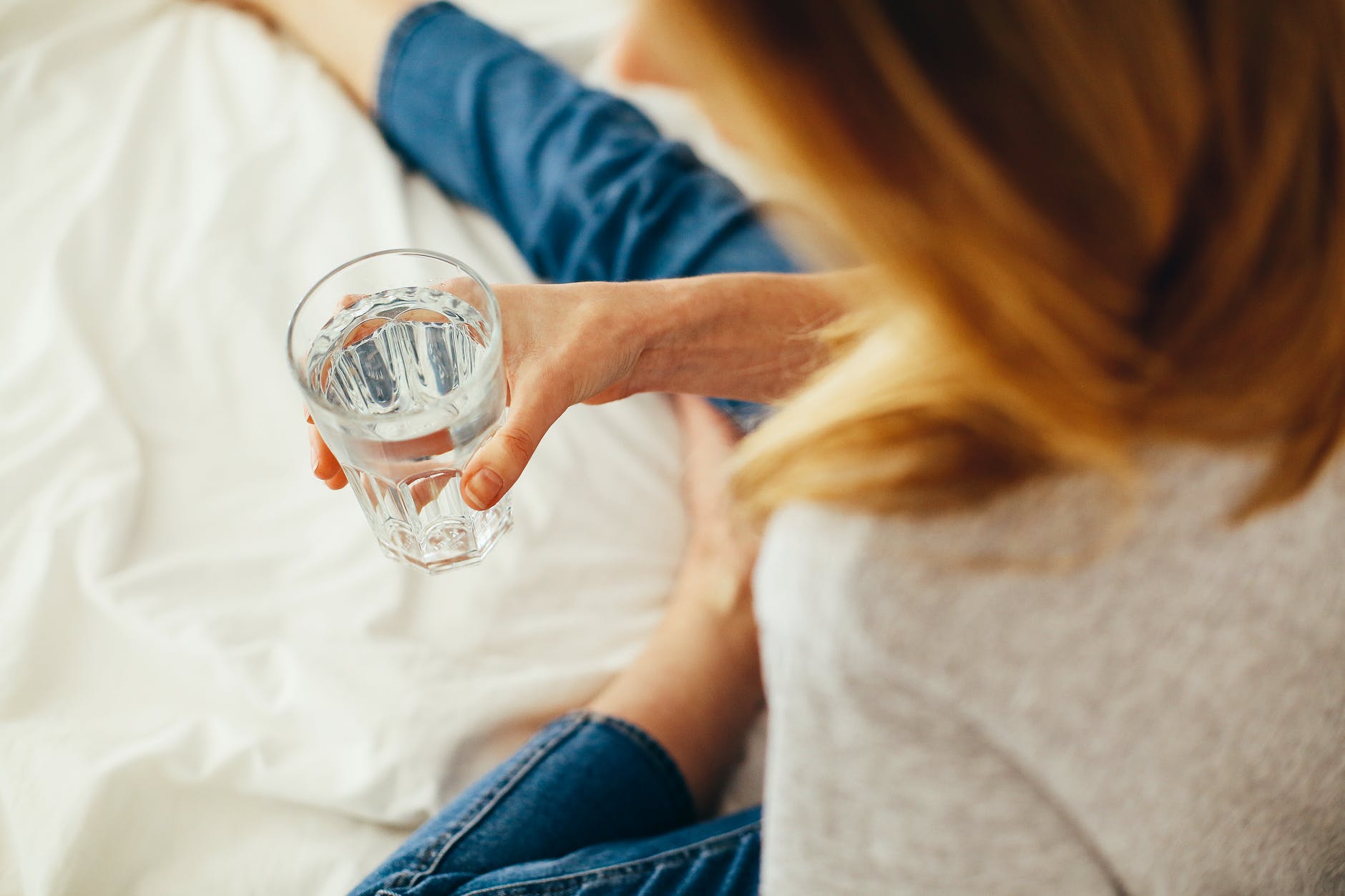 Una mujer sentada en una cama sosteniendo un vaso de vidrio lleno de agua. | Foto: Pexels
