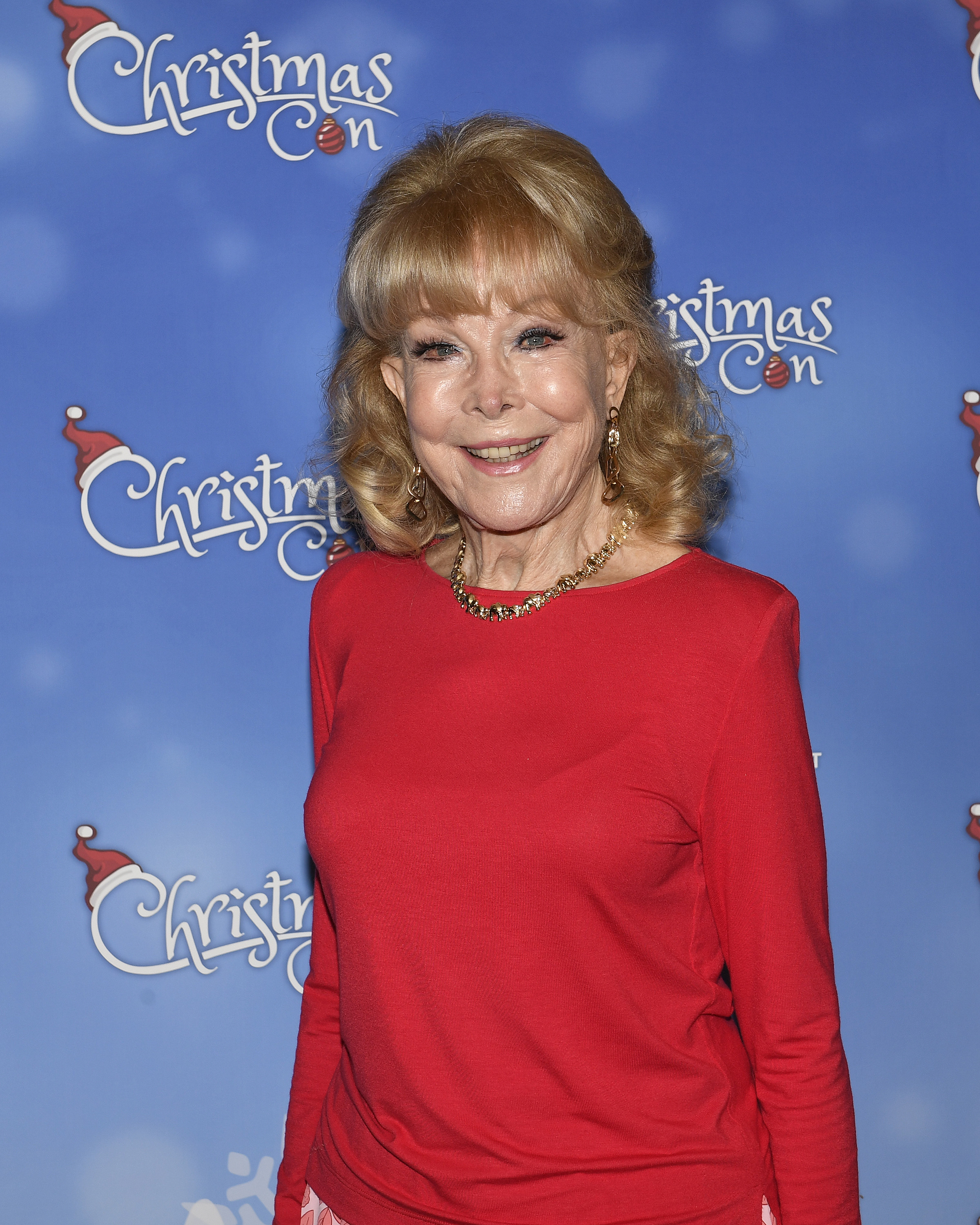 Barbara Eden smiles on the blue carpet at Christmas Con in Pasadena, wearing a bright red top, gold necklace, and softly curled blond hair.