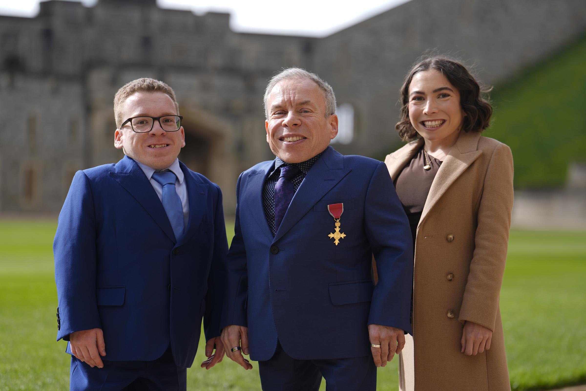 Warwick Davis is flanked by his son, Harrison Davis, 23, and daughter, Annabelle Davis, 28, on the grounds of Windsor Castle on 11 March 2026, all three grinning broadly for the camera with the castle's ancient stone walls stretching out behind them. It is a family portrait with real weight to it — the children who stood by their father through grief were also there to witness one of the proudest moments of his career.