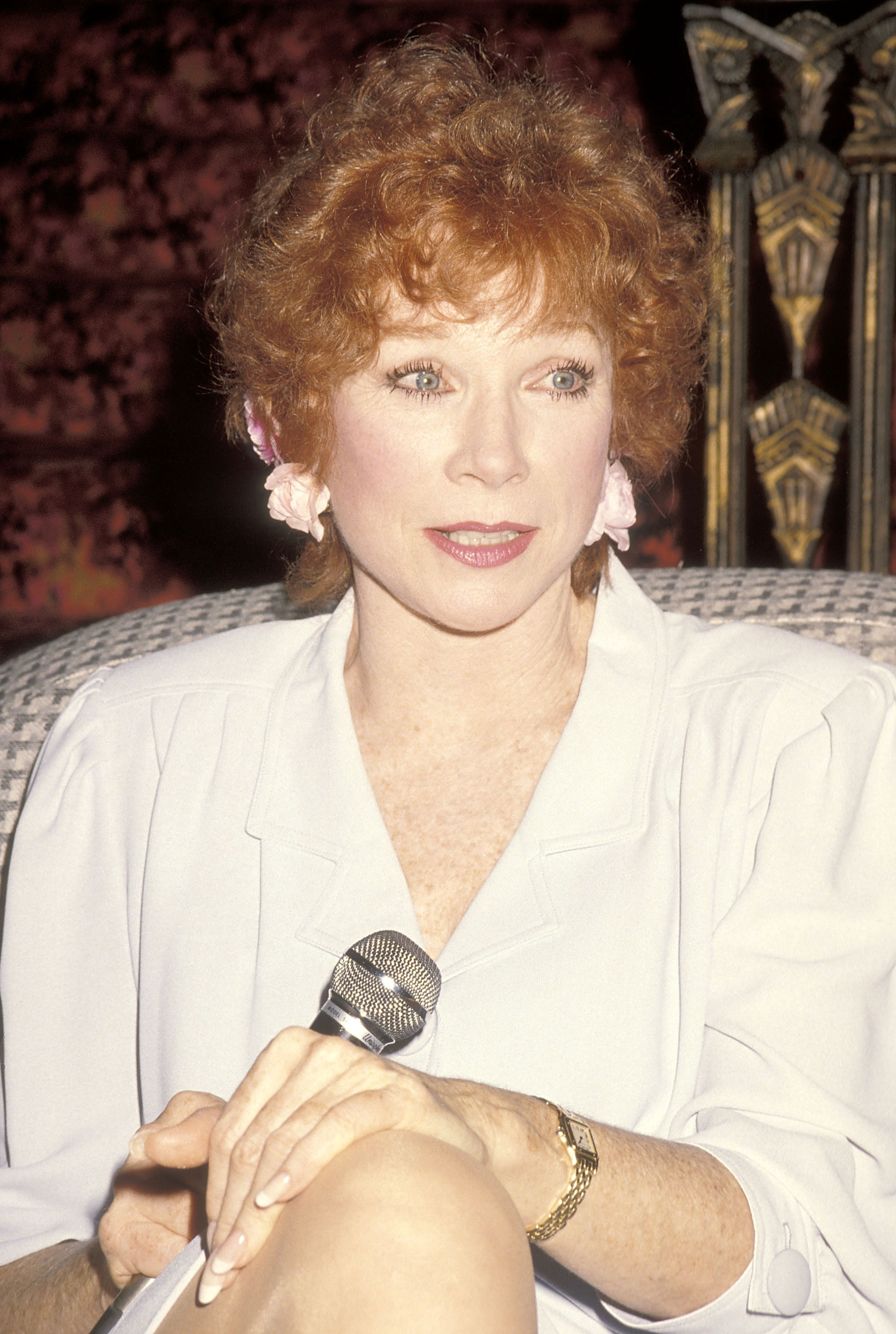 Poised and pensive, MacLaine speaks at the 1990 "Out There Tonight" press conference at the Pantages Theatre. Dressed in a soft white look and holding the mic with quiet strength, she reflects on her personal journey and powerful stage return.