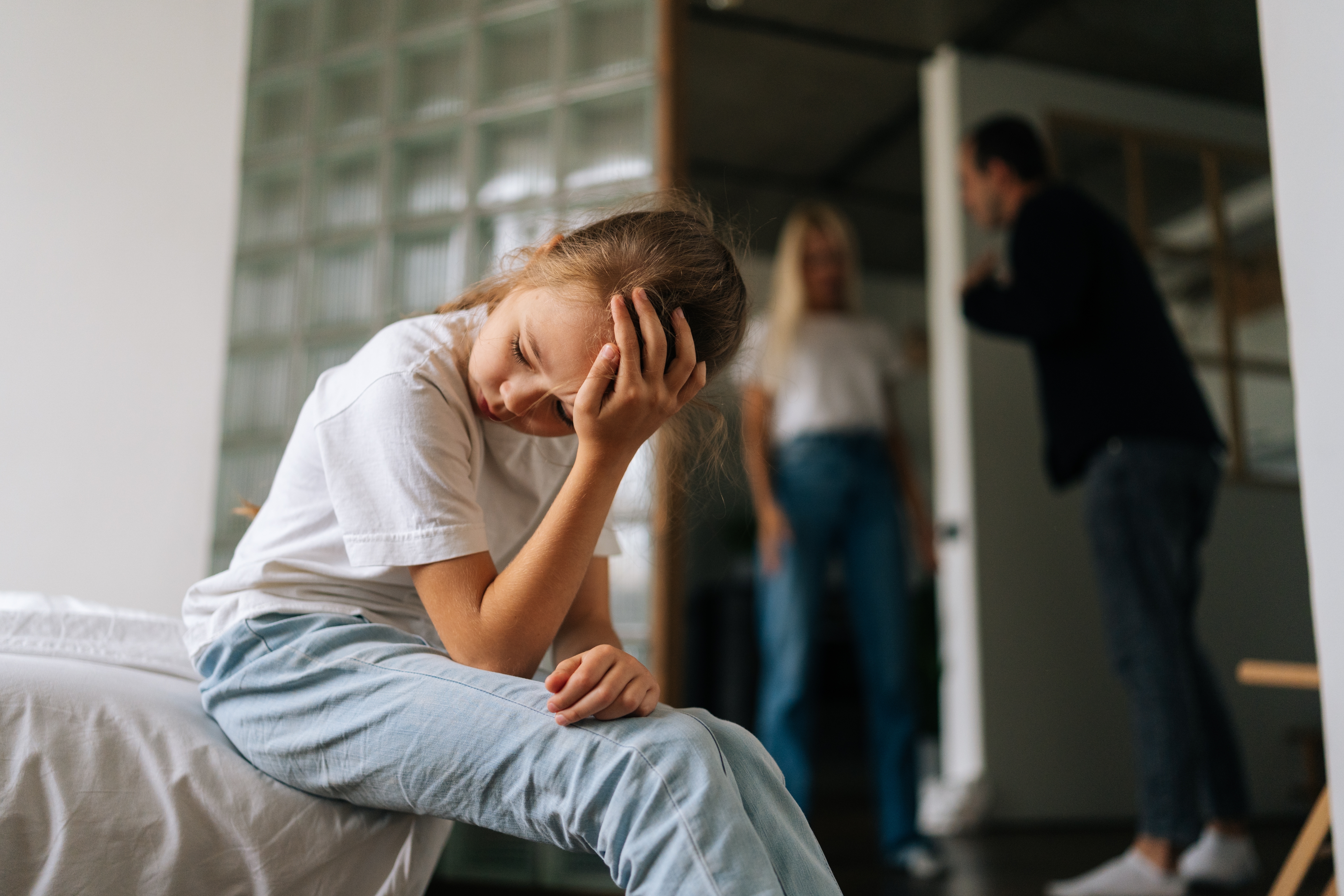 An upset girl sitting on the corner of her bed while her parents argue in the background | Source: Shutterstock