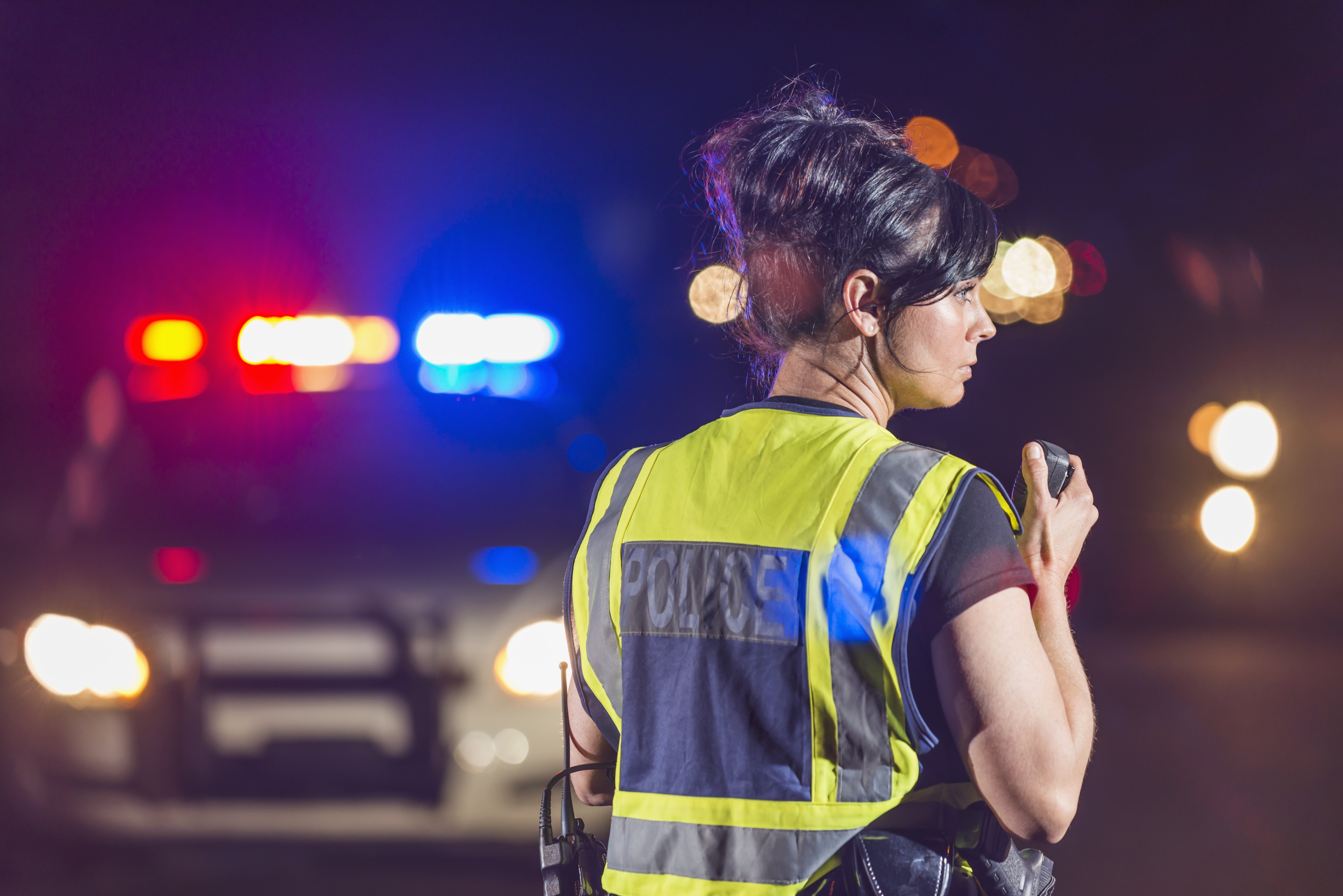 Rear view of a female police officer standing in the street at night, talking into her radio. Her patrol car is in the background with the emergency lights illuminated. She is wearing a yellow safety vest|Photo: Getty Images
