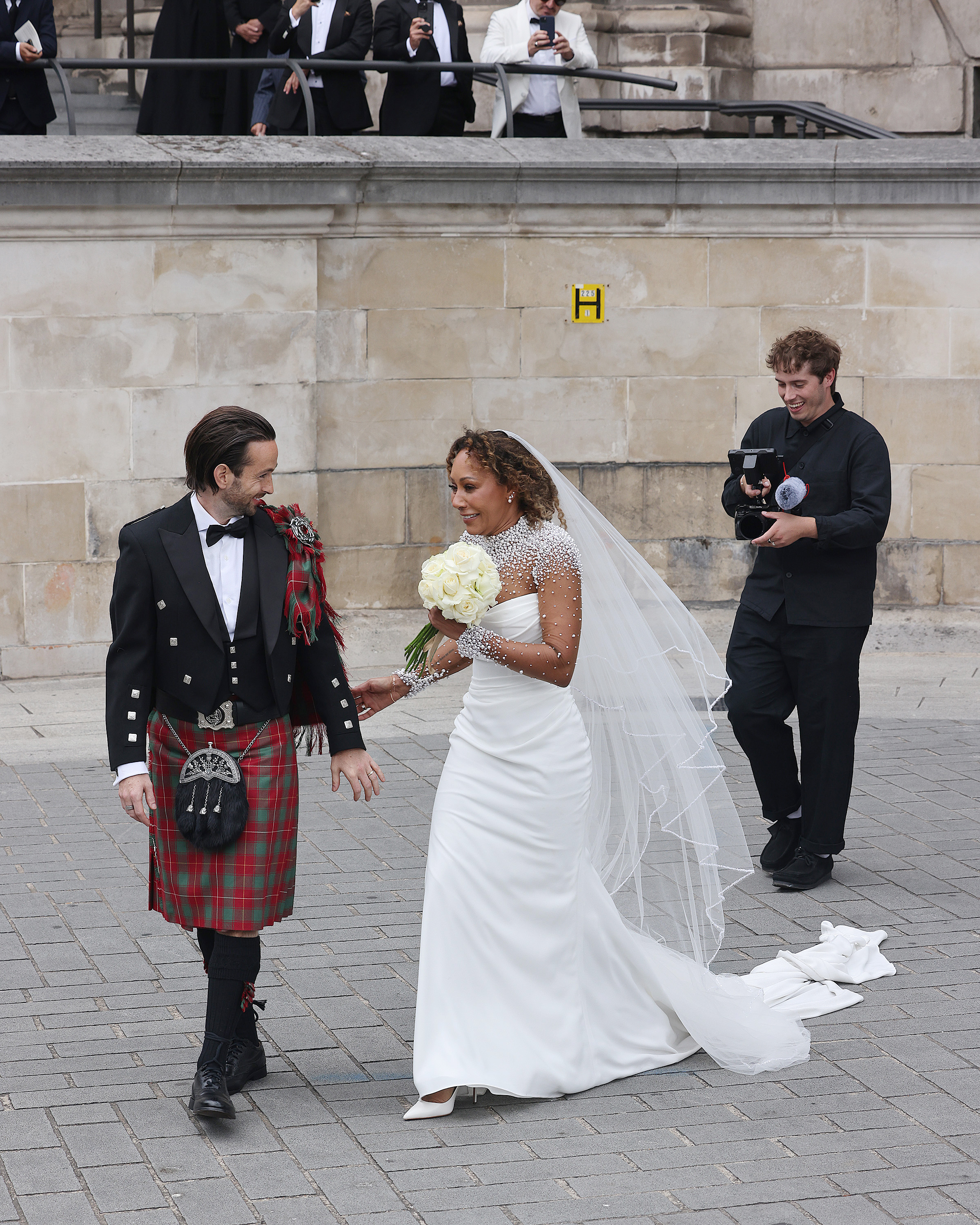 As they walk away from the cathedral, Mel B and Rory McPhee smile warmly, their backs to the camera. A photographer trails behind, capturing the newlyweds from a distance. The image offers a graceful, almost cinematic close to their ceremony, filled with quiet joy and soft movement.