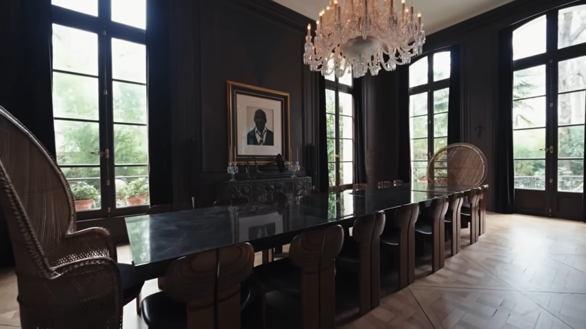 High arched windows frame the moody dining room, where low-light ambiance and rich wood textures create a warm, welcoming space for dinner parties. | Source: YouTube/@Archdigest