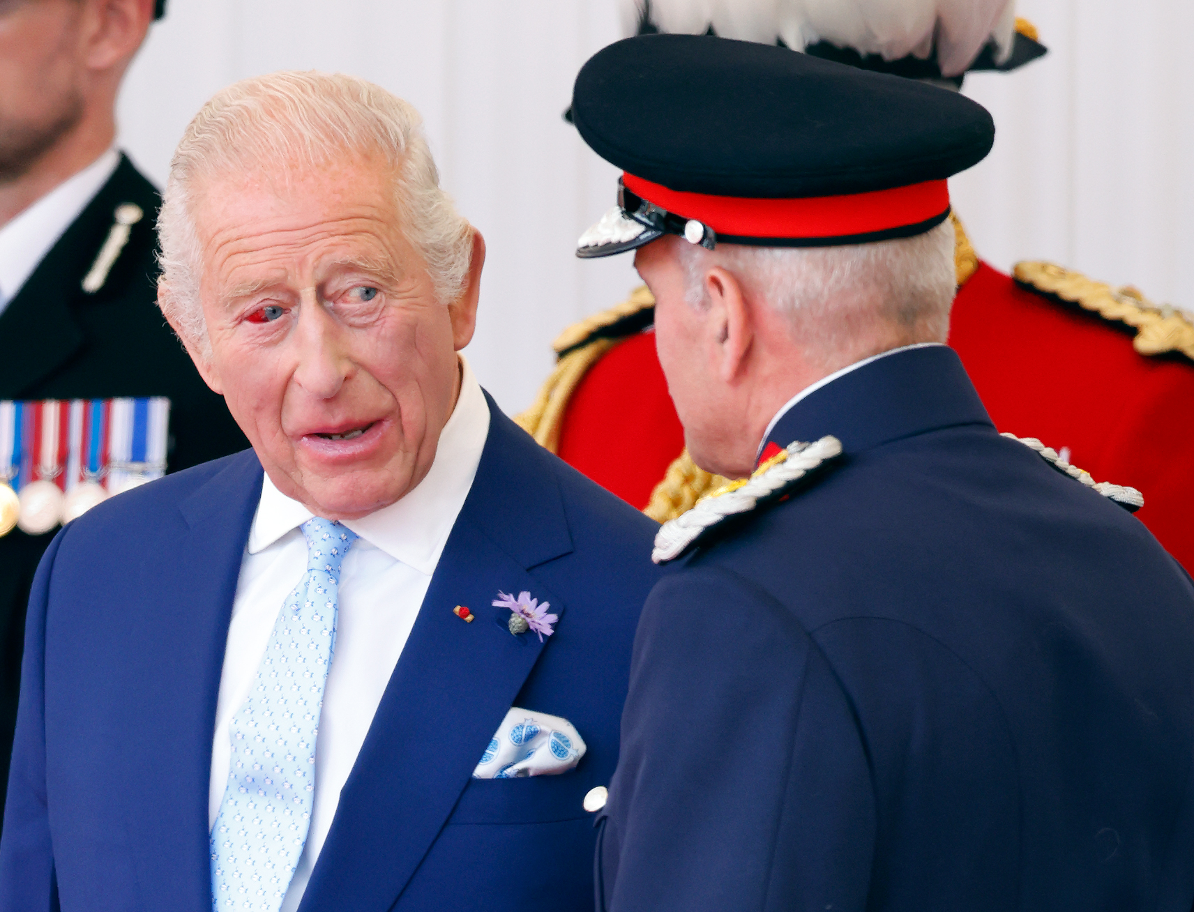 A close-up of King Charles III reveals the red tint in his right eye as he converses with a man in uniform. Despite the burst vessel, the king remains engaged throughout the official welcoming.