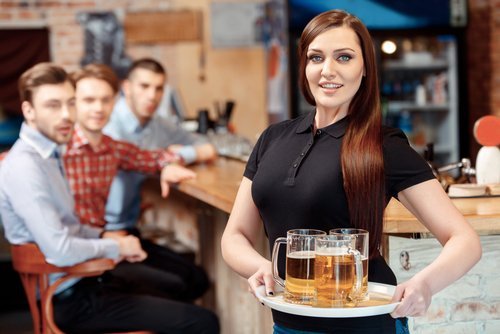 Une belle serveuse tenant un plateau avec des verres à bière pendant que les jeunes hommes la regardent avec étonnement | Photo : Shutterstock Une belle serveuse tenant un plateau avec des verres à bière pendant que les jeunes hommes la regardent avec étonnement | Photo : Shutterstock