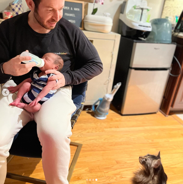 A photo of Brandon Frankel feeding his son, Cooper, a bottle of milk while his cat looks on, posted in June 2024 | Source: Instagram/gabby3shabby