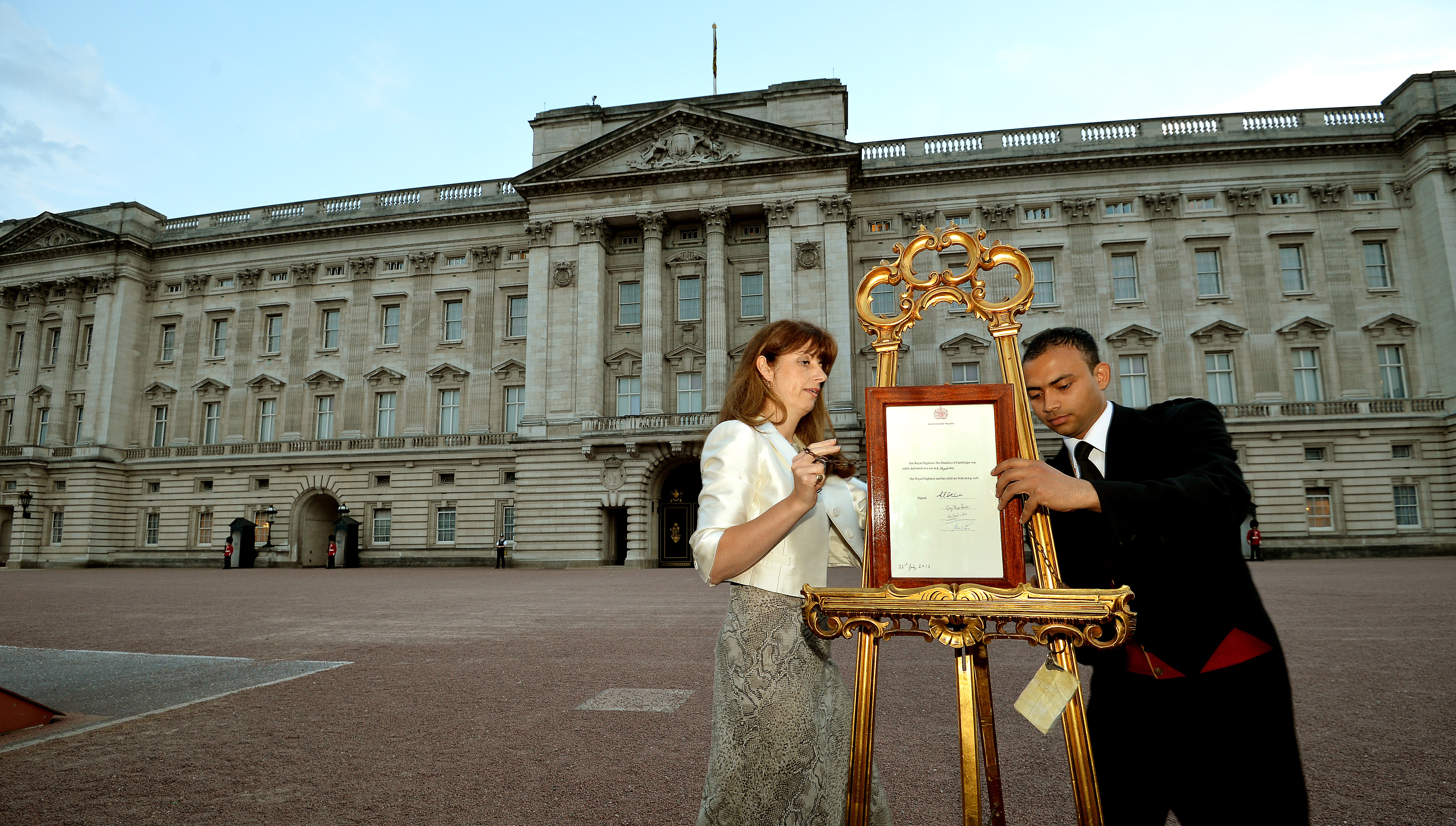 Ailsa Anderson with Badar Azim, a footman, place on an easel in the forecourt of Buckingham Palace a notification to announce the birth of Prince George on July 22, 2013 | Source: Getty Images