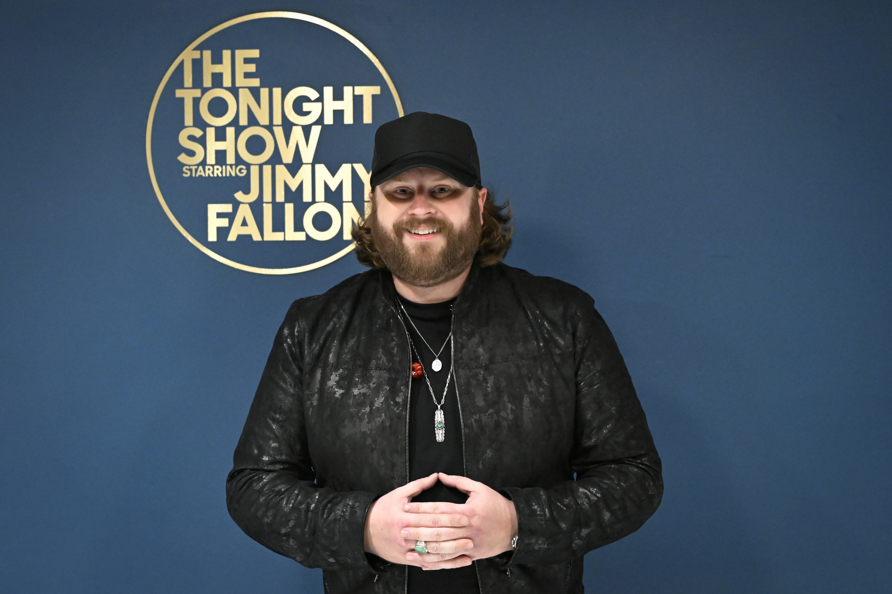 Nate Smith poses backstage at “The Tonight Show Starring Jimmy Fallon,” smiling in an all-black ensemble complete with a cap, layered necklaces, and a patterned jacket.