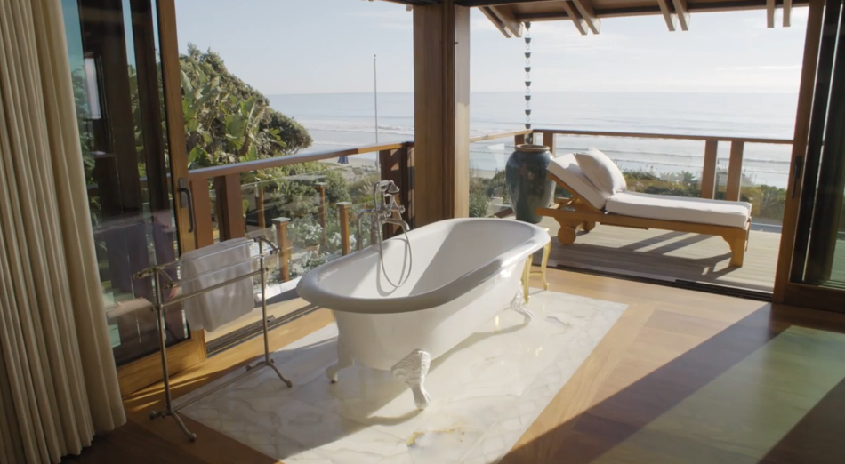 Set on an inset marble tile floor, a classic white clawfoot soaking tub with polished chrome fixtures takes center stage near expansive glass doors. The doors retract completely to connect the warm hardwood floors of the bathroom to a private upper deck equipped with a lounge chair overlooking the Pacific Ocean. | Source: YouTube/Architectural Digest