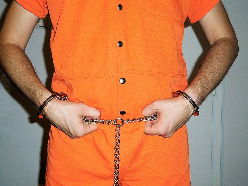 Primer plano de un recluso en bata institucional naranja y restricciones de transporte de "arnés completo". | Imagen: Wikimedia Commons Primer plano de un recluso en bata institucional naranja y restricciones de transporte de "arnés completo". | Imagen: Wikimedia Commons