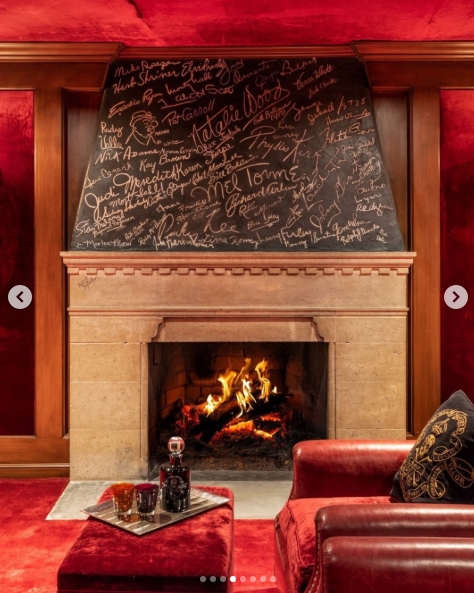 The home theatre fireplace that became a piece of living history: a blackboard above the mantel is covered edge to edge in the signatures of those who passed through these rooms, including Natalie Wood and Mel Tormé, their names visible amongst dozens of others. A log fire burns below, and the room's deep red walls and leather seating give it the feeling of a private members' club. | Source: Instagram/top.music.songs
