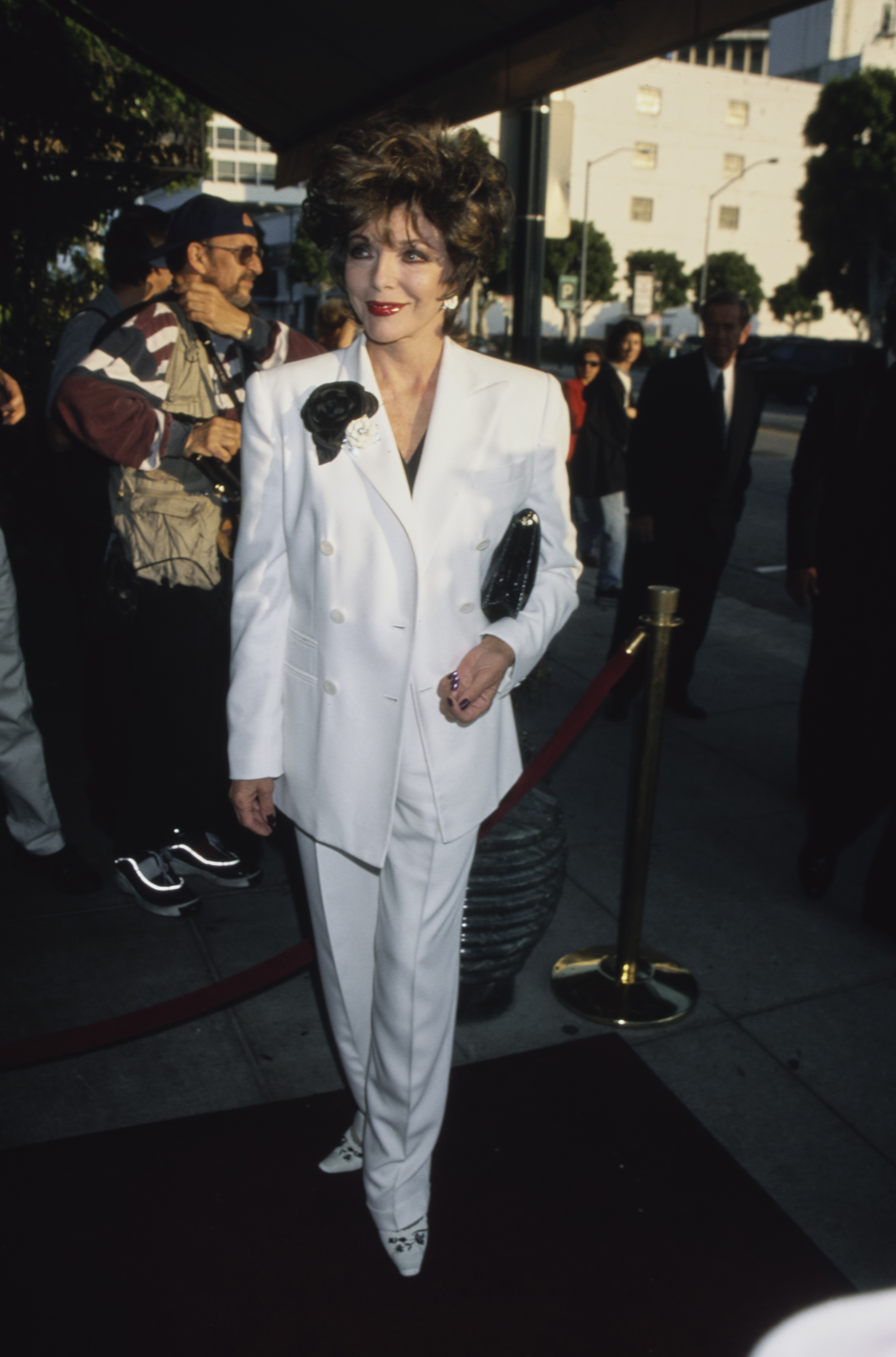 Evergreen Creator said: Joan Collins arrived at the Beverly Hilton Hotel on June 7, 1999, for the R.A.D.D. Benefit Gala honouring Liza Minnelli, dressed in a crisp white double-breasted suit adorned with a dramatic black flower brooch. Her structured tailoring, glossy clutch, and signature voluminous hair captured a refined take on late-1990s red carpet elegance.