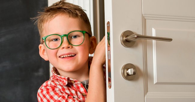 A little boy with green glass peeps around a white door. | Photo: Shutterstock