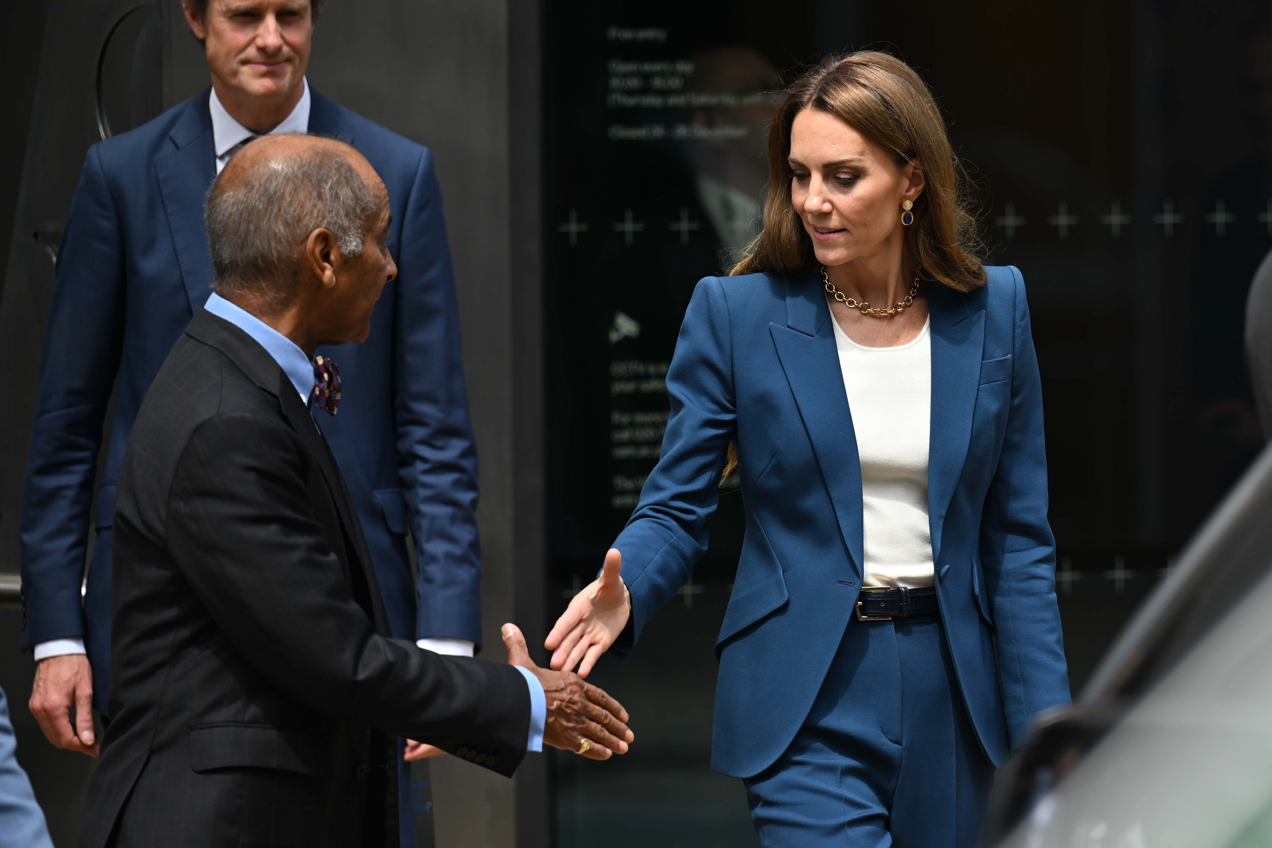 Princess Catherine reaches out for a handshake with a calm, focused look. Her tailored blue suit and composed stance convey elegance and attentiveness in a formal setting.
