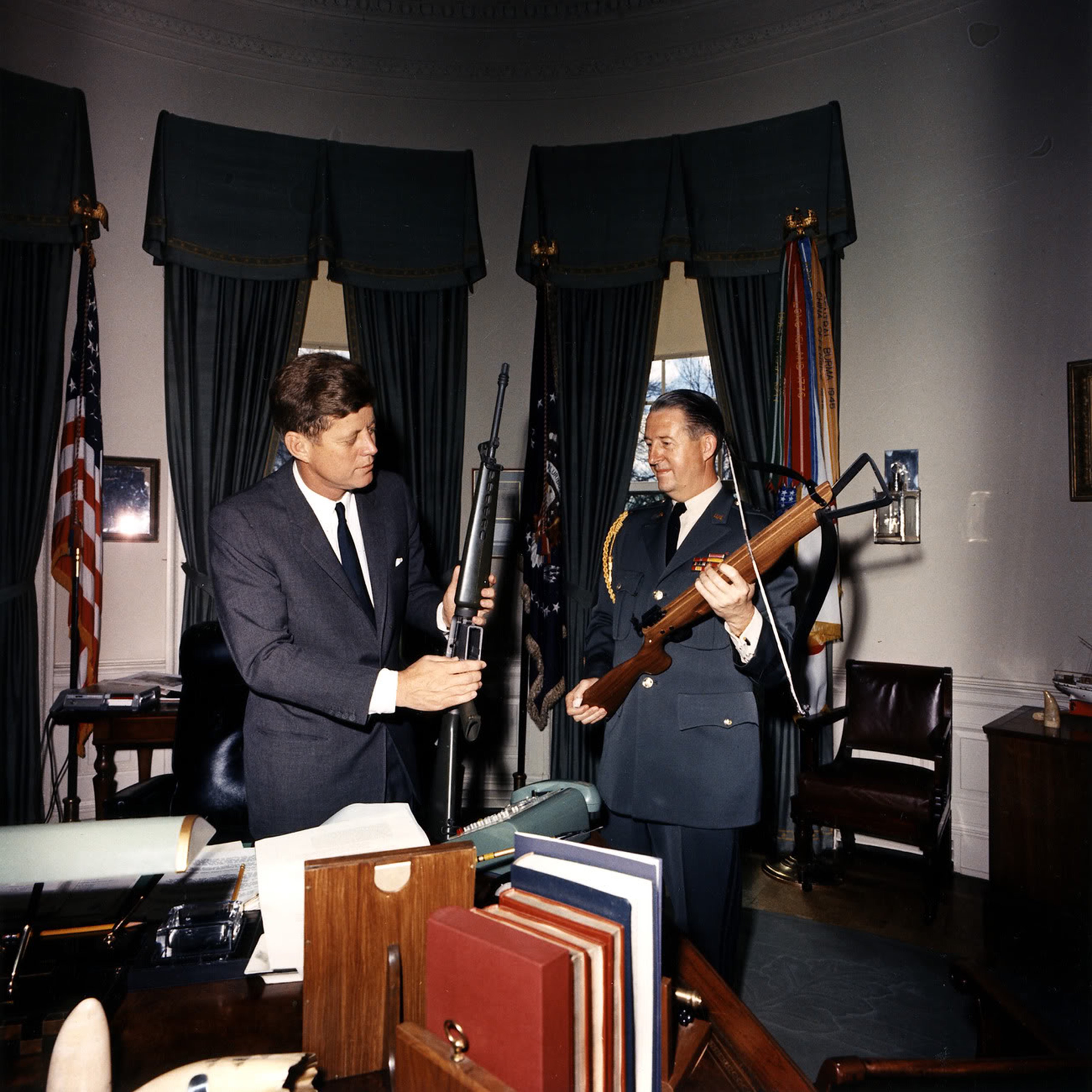 On April 19, 1963, President John F. Kennedy inspects a rifle beside Major General Ted Clifton in the Oval Office, where dark drapes, military flags, and a cluttered desk reflect the era's décor.