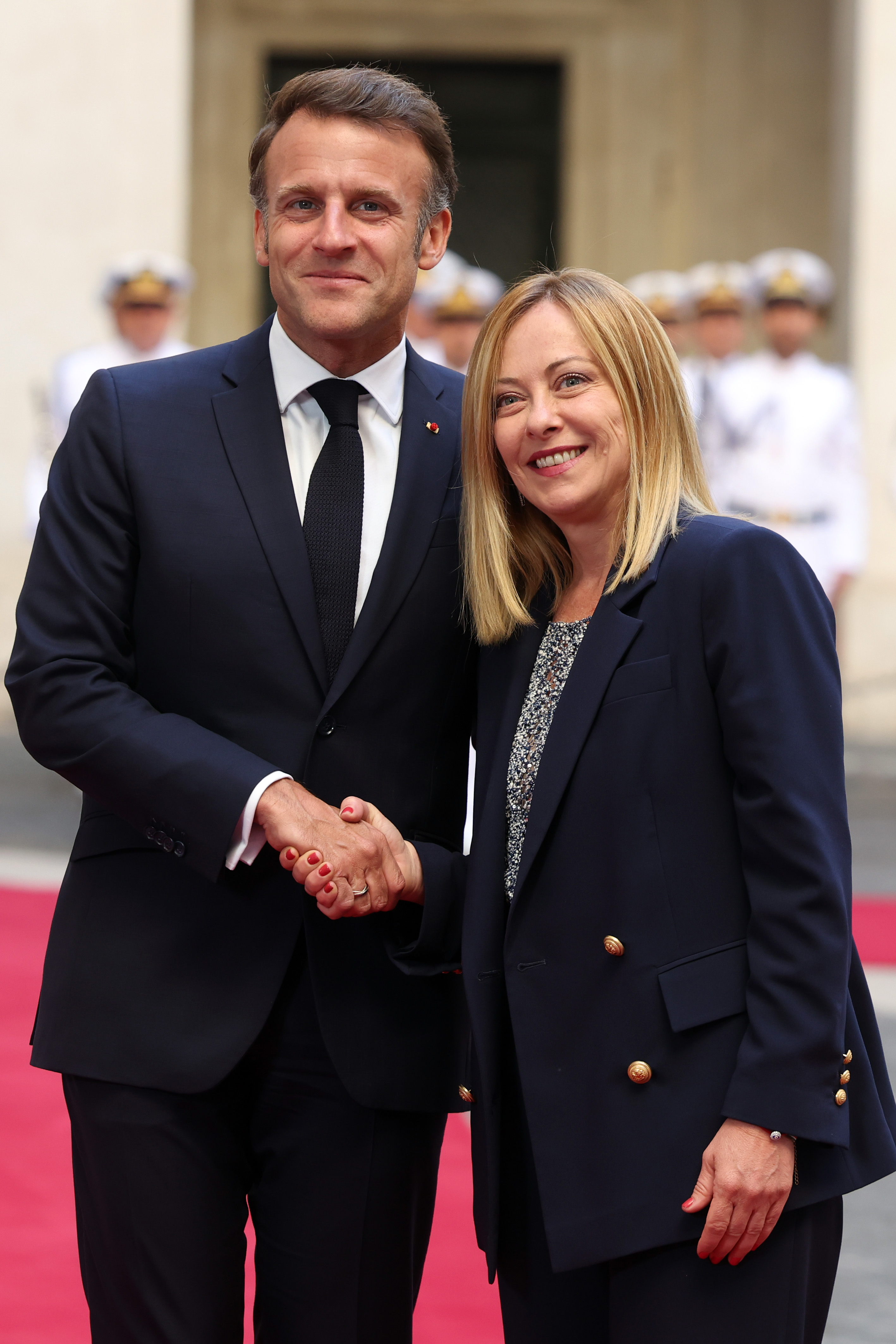 Emmanuel Macron and Giorgia Meloni pose side by side outside Palazzo Chigi, smiling warmly as they shake hands. The moment captures a formal yet friendly exchange between the two leaders.