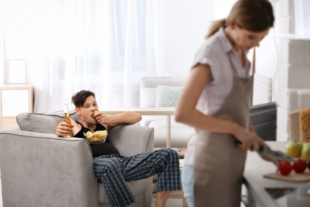 A lazy husband sits on a couch and has a snack while watching TV as his wife slaves away cooking in the kitchen | Photo: Shutterstock/New Africa