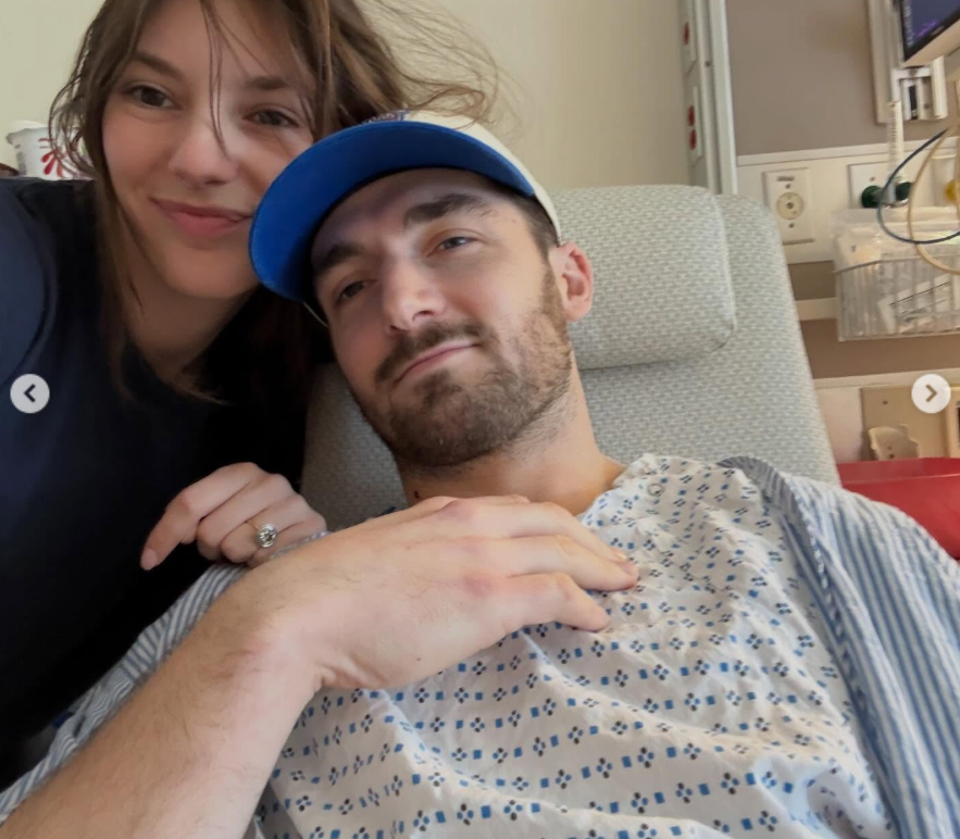Natalie Ackerman leans close beside Daniel Neeson as they pose for a selfie in a hospital chair. He wears a hospital gown and cap while she rests a hand on his shoulder, her ring visible. | Source: Instagram/bgdans91