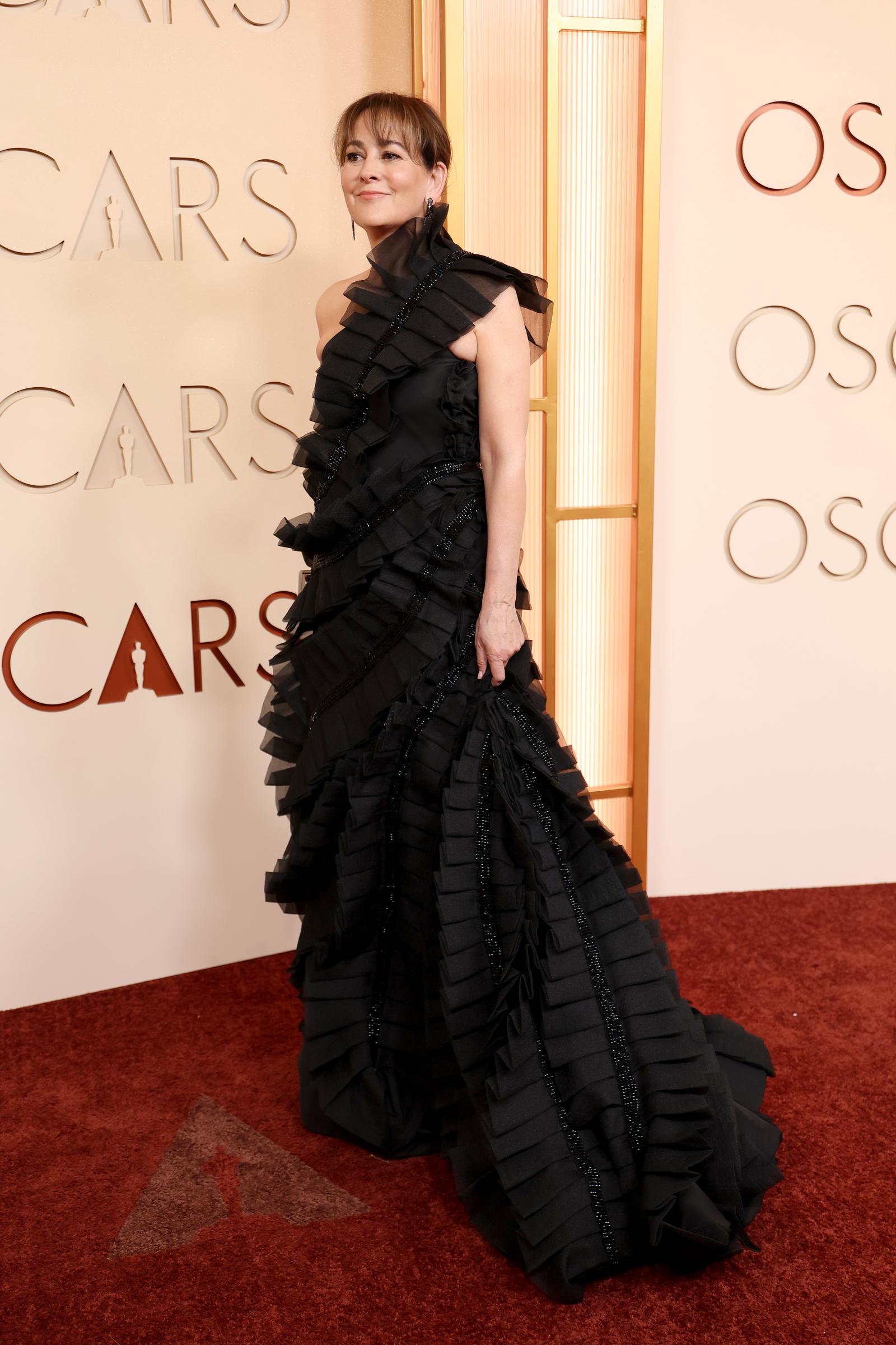 Lesley Barber is photographed side-on at the ceremony in a voluminous all-black ballgown covered from shoulder to hem in tightly layered horizontal ruffled fabric panels, with an asymmetric one-shoulder neckline and a sheer ruffle at the collar sweeping into a full trailing skirt.