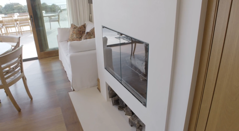 A sleek glass-enclosed fireplace built into a white stucco wall adds modern warmth beside a plush white loveseat outfitted with patterned accent pillows. Beneath the glass hearth, integrated cubbies keep neatly stacked firewood ready for use right near the dining area. | Source: YouTube/Architectural Digest