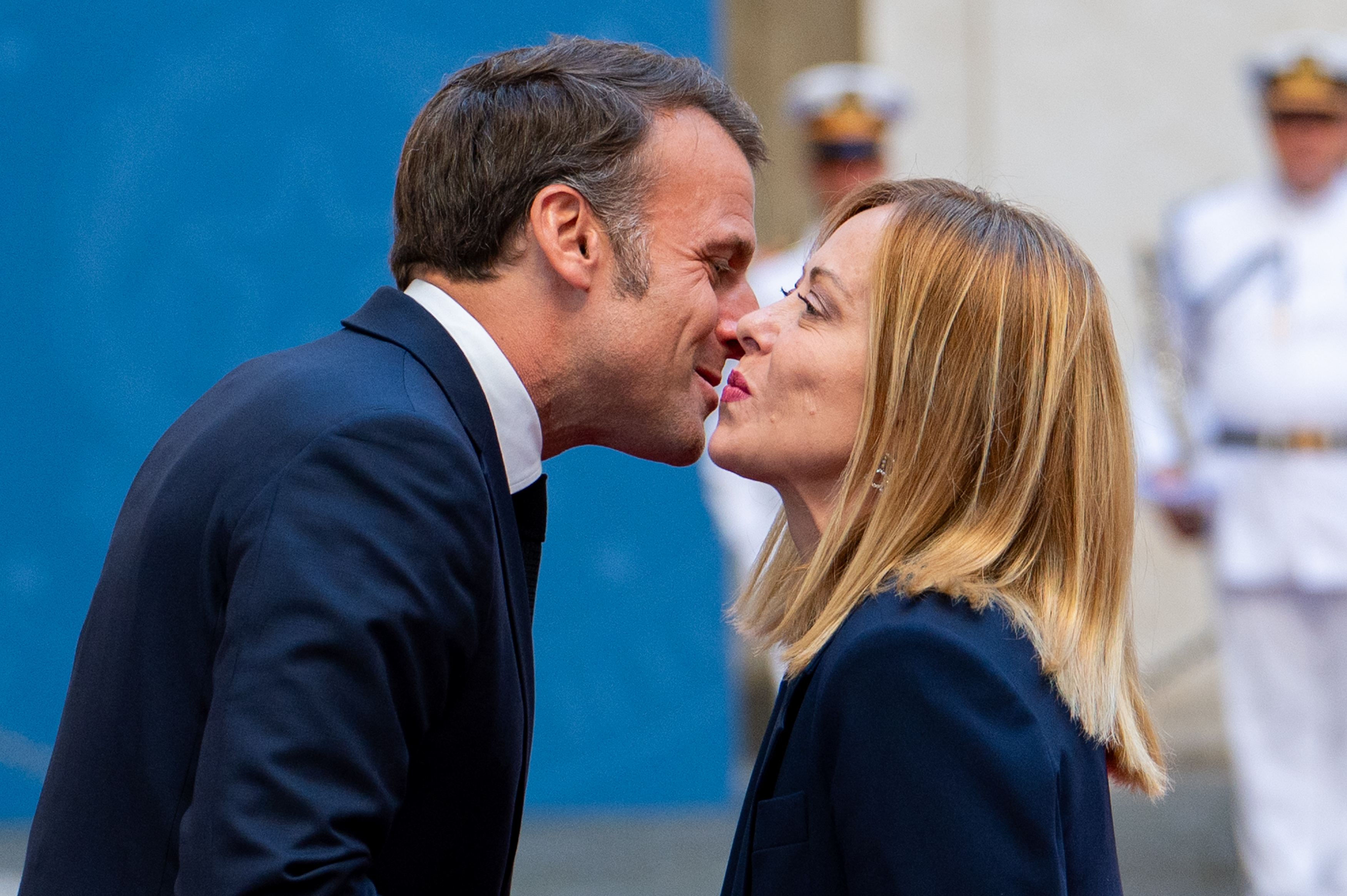 Emmanuel Macron leans in to greet Giorgia Meloni with a traditional cheek kiss as they stand closely face to face. Both leaders maintain direct eye contact, with uniformed personnel blurred in the background.