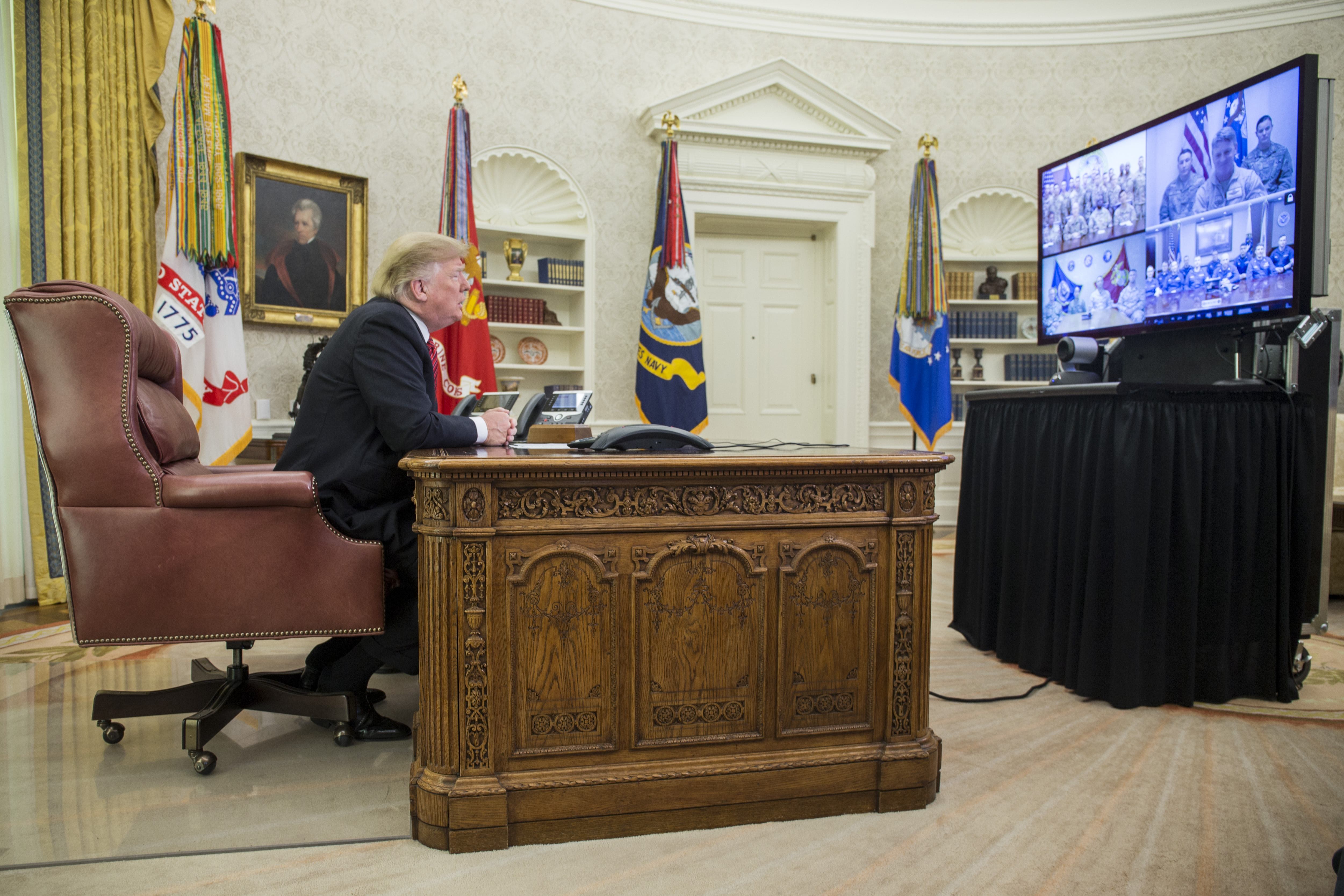 President Donald Trump speaks with U.S. troops on December 25, 2018, from the Oval Office, featuring the ornate Resolute Desk, presidential flags, and historic portraits.