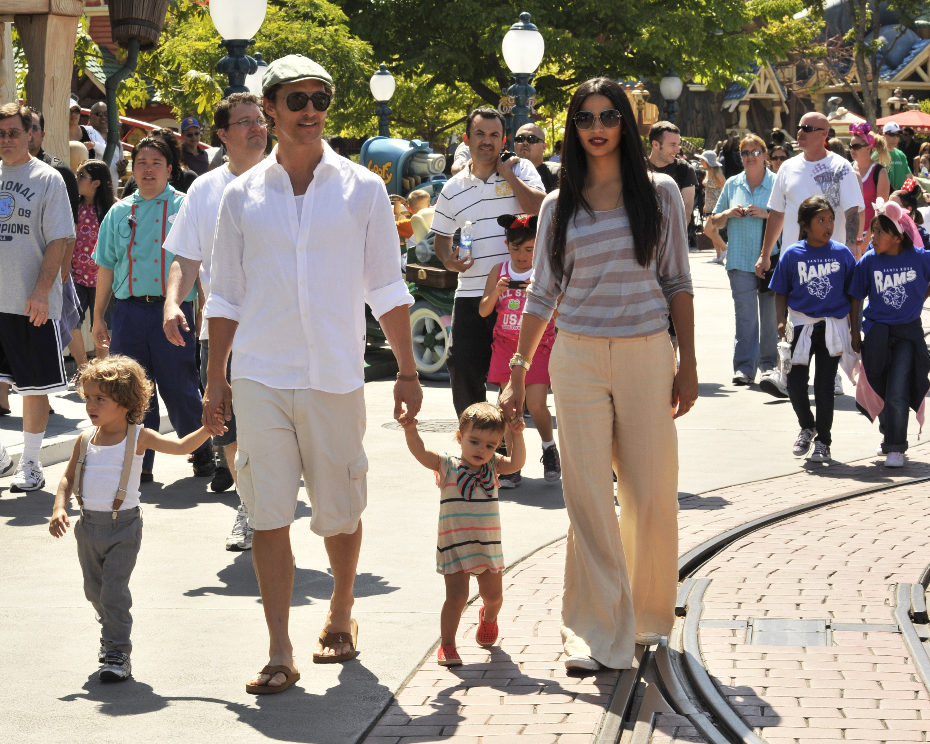 Levi McConaughey and Vida McConaughey walk between Matthew McConaughey and Camila Alves. They wear light, casual outfits suited for a day outdoors. Matthew McConaughey and Camila Alves walk alongside them through the park.