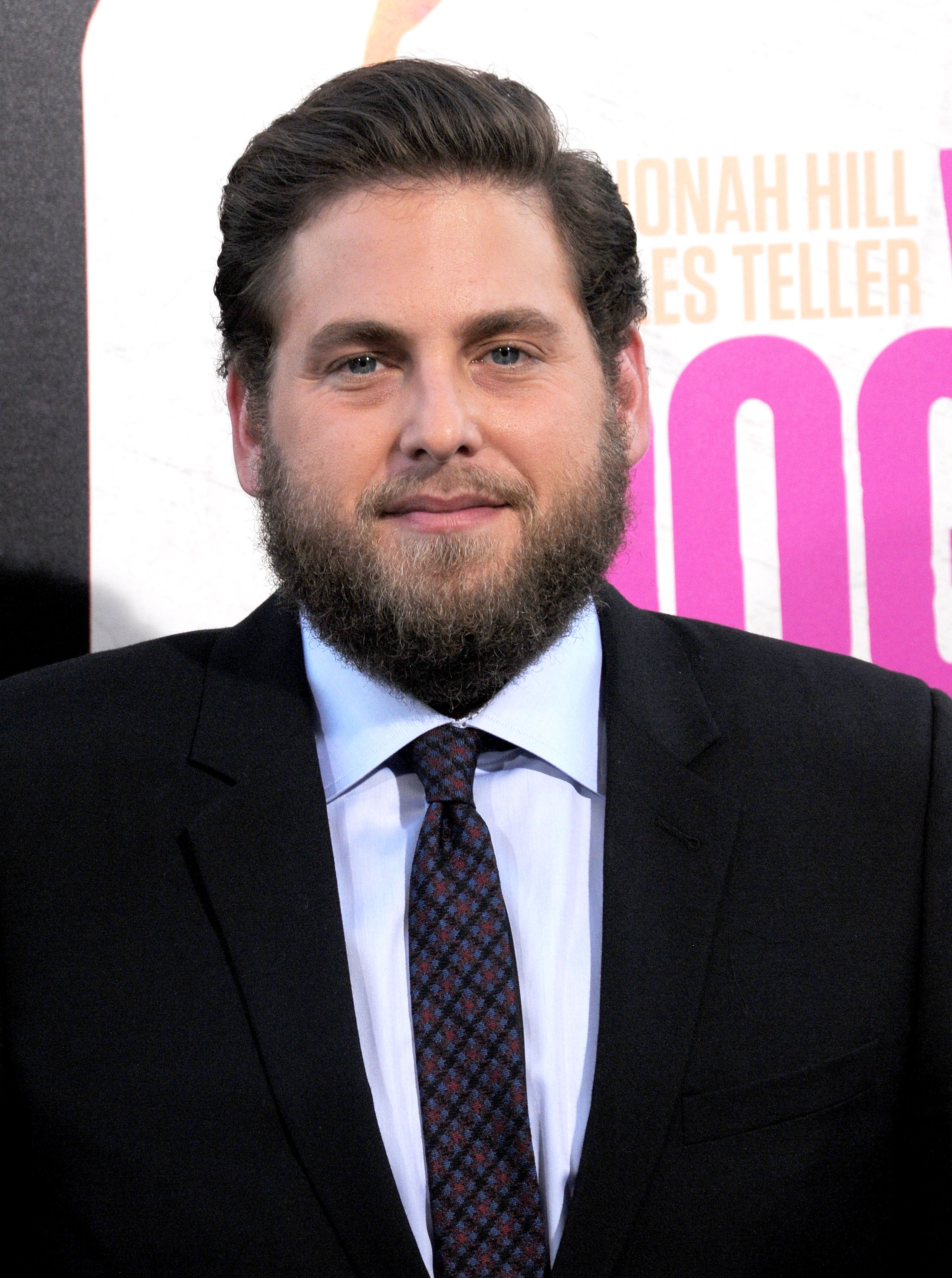 Jonah Hill poses at the premiere of 'War Dogs' in a black suit and patterned tie, his full beard and slicked-back hair giving him a more rugged look. The actor stands in front of a promotional backdrop featuring his name in bold letters.