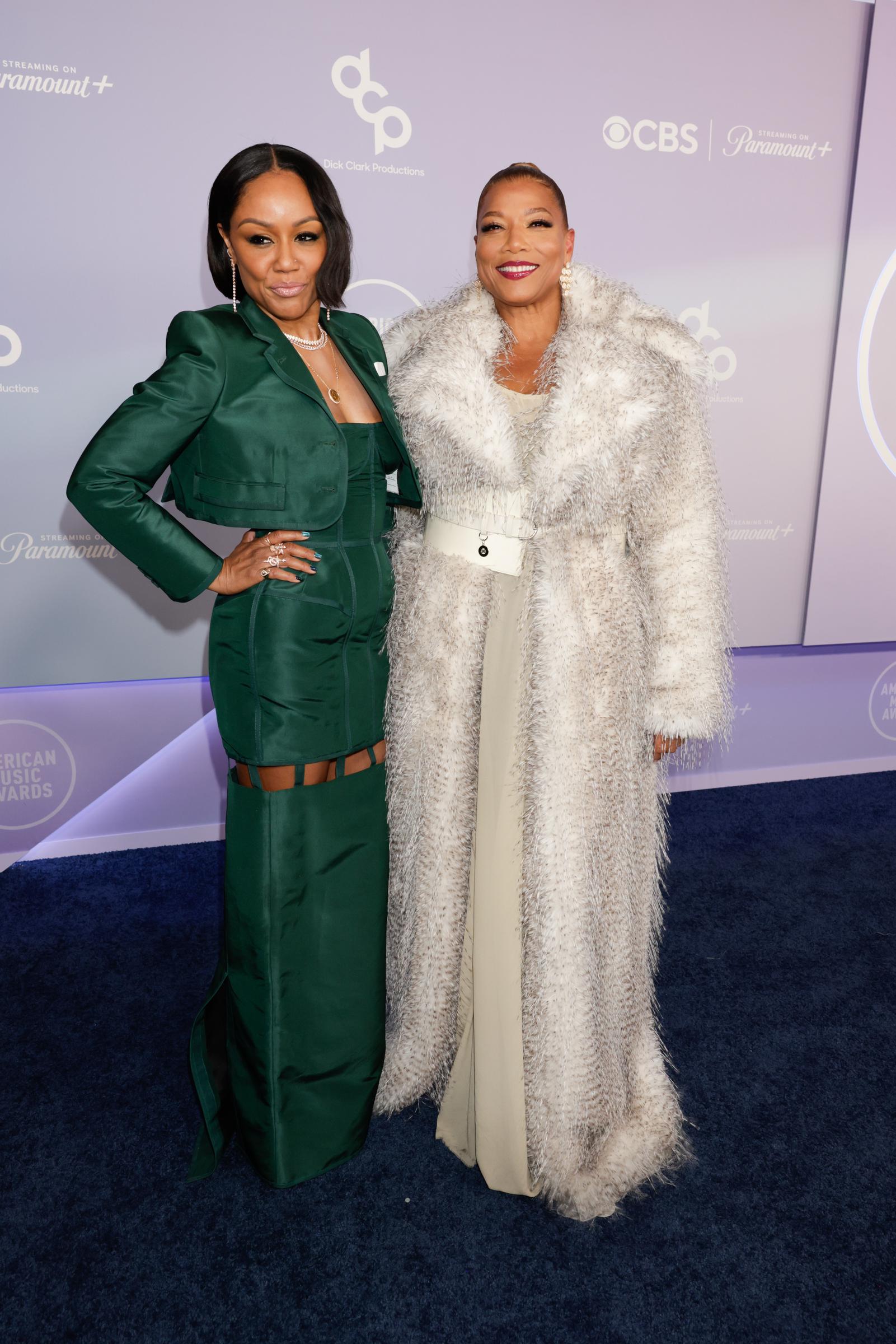 Eboni Nichols and Queen Latifah stand side by side on the AMAs carpet, showcasing coordinated glamour in structured emerald green and shimmering silver looks under bright lights.