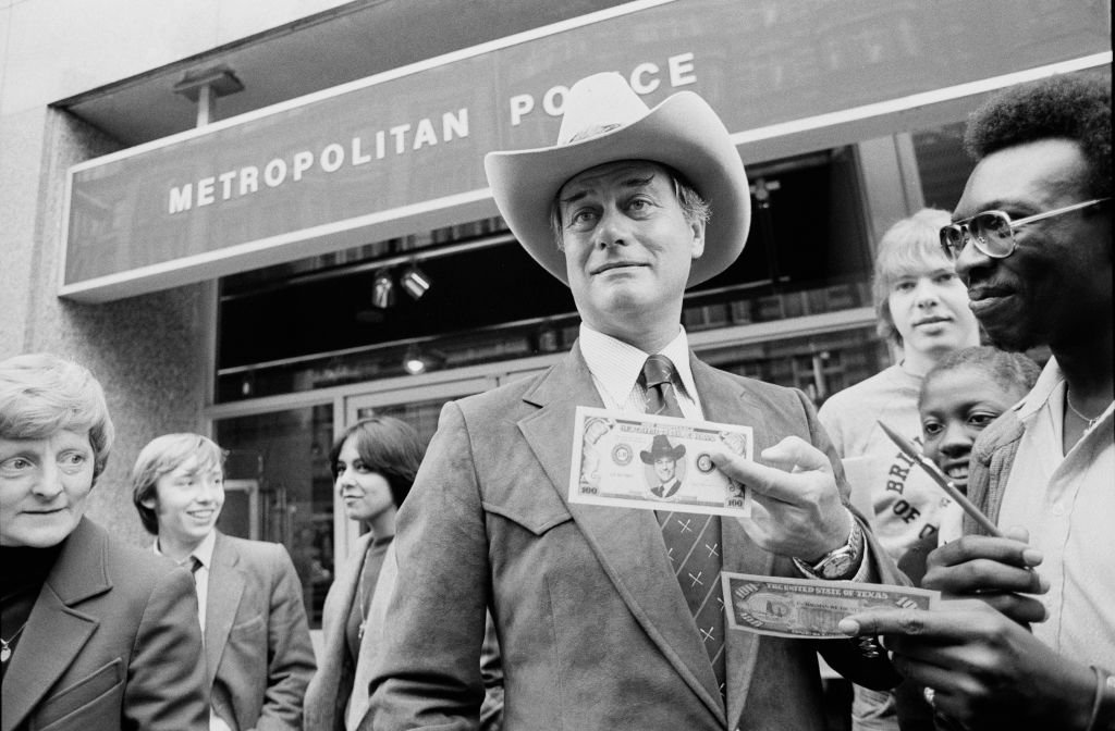 Larry Hagman (1931 - 2012) signing $100 bill from television show Dallas, in which he plays 'JR Ewing', outside the Metropolitan Police offices, London, UK, 10th June 1980. | Source: Getty Images