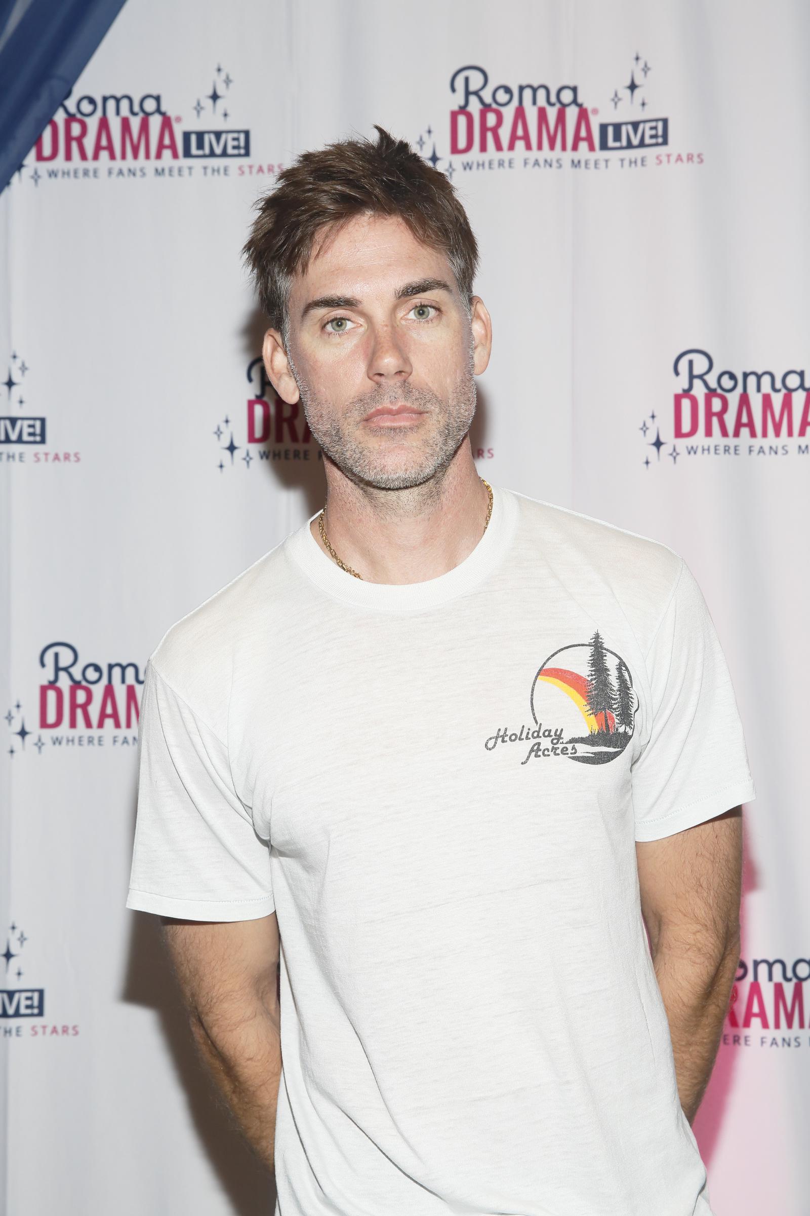 Drew Fuller attends RomaDrama Live! in 2022, dressed casually in a white "Holiday Acres" graphic T-shirt. With a relaxed stance and subtle stubble, he gazes directly into the camera against a branded step-and-repeat backdrop. The laid-back look reflects the fan-focused atmosphere of the event.