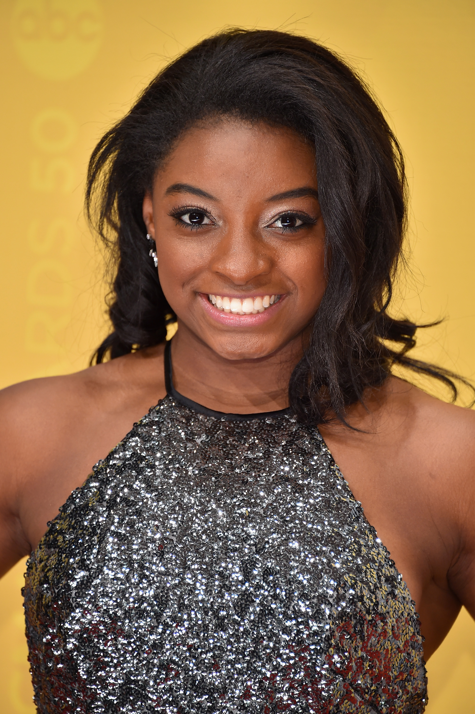 Dressed for the 50th annual CMA Awards, Simone Biles smiles brightly against a bold yellow background. Her hair is styled in soft waves, framing her face with effortless elegance. The look blends youthful energy with refined glamour.