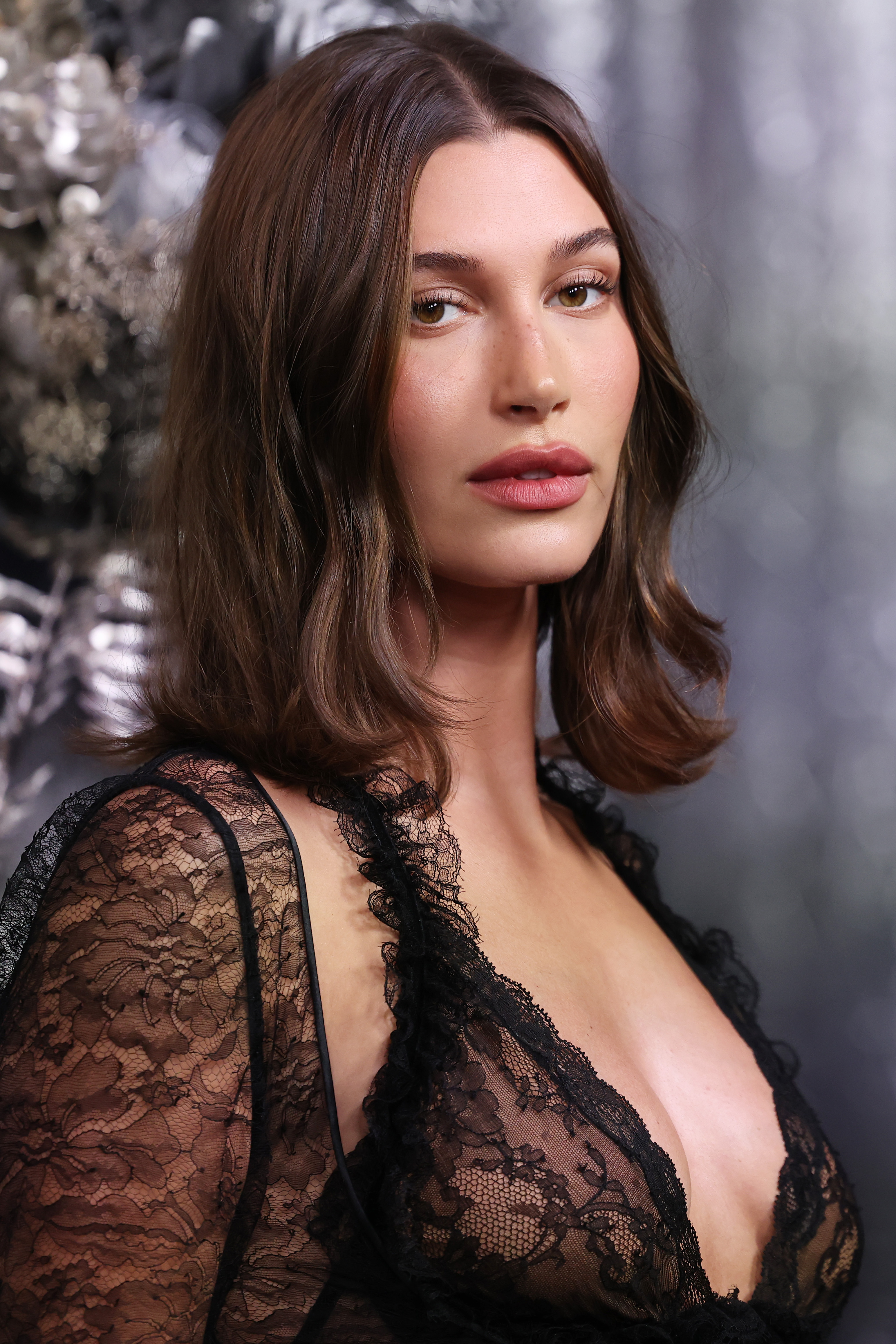 Hailey Bieber captivated in a sheer black lace gown with a plunging neckline, her soft brunette hair and natural glam makeup enhancing the sultry, refined aesthetic.